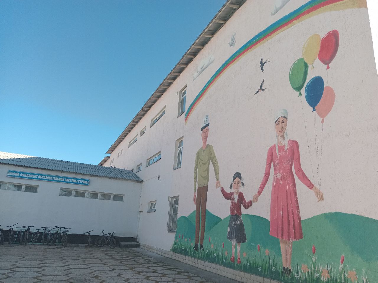 mural on a white school building of a man wearing a white Kyrgyz hat, a little girl, and a woman holding hands. balloons in the woman's second hand and a couple birds and a rainbow above the people
