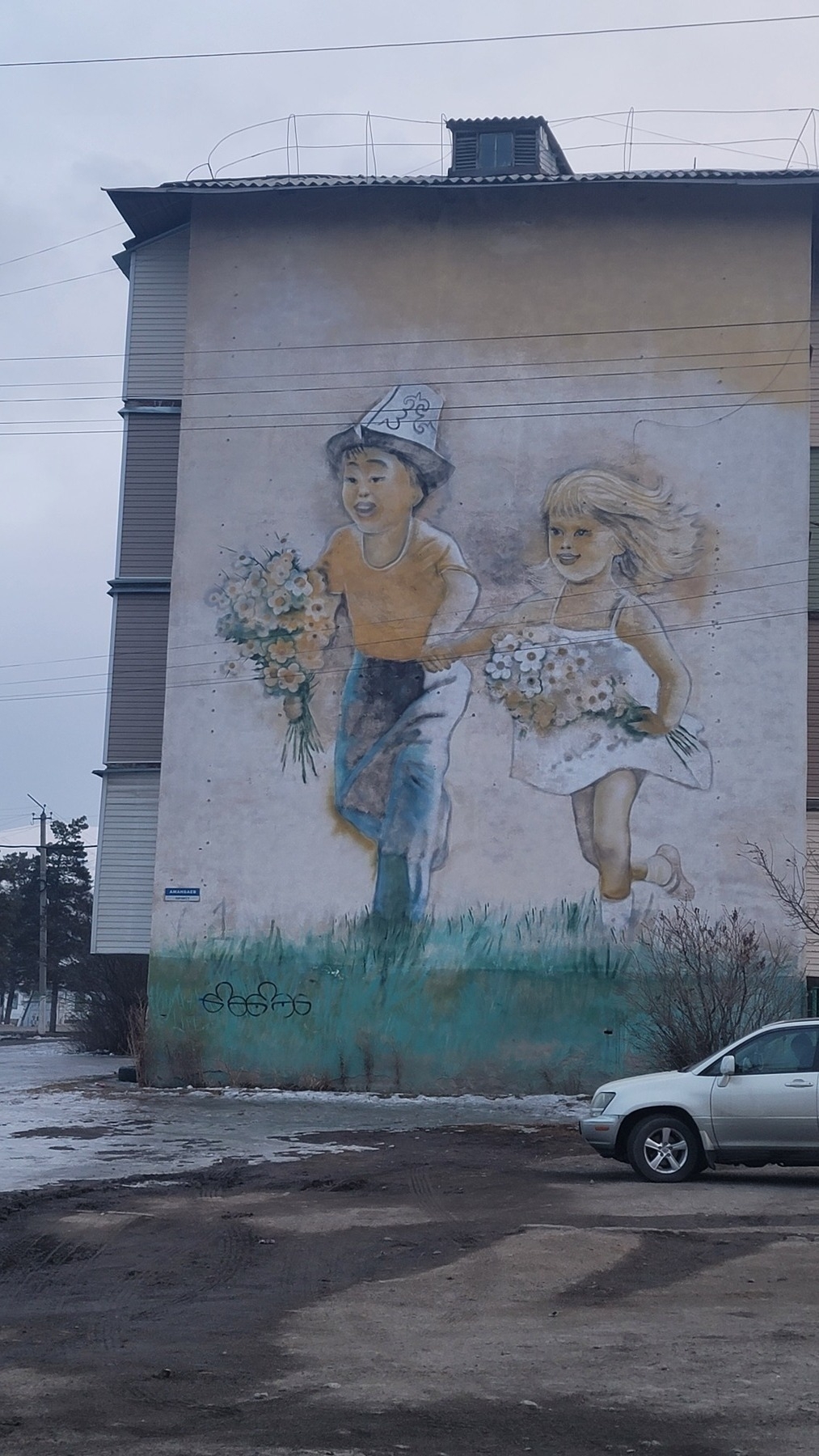 mural on a tan wall of a 4-story building with a young Kyrgyz boy wearing a Kyrgyz hat, orange t-shirt, and jeans; and a little pale, blonde girl wearing a white strap dress. the two children are holding hands and both holding a bunch of flowers, skipping through grass