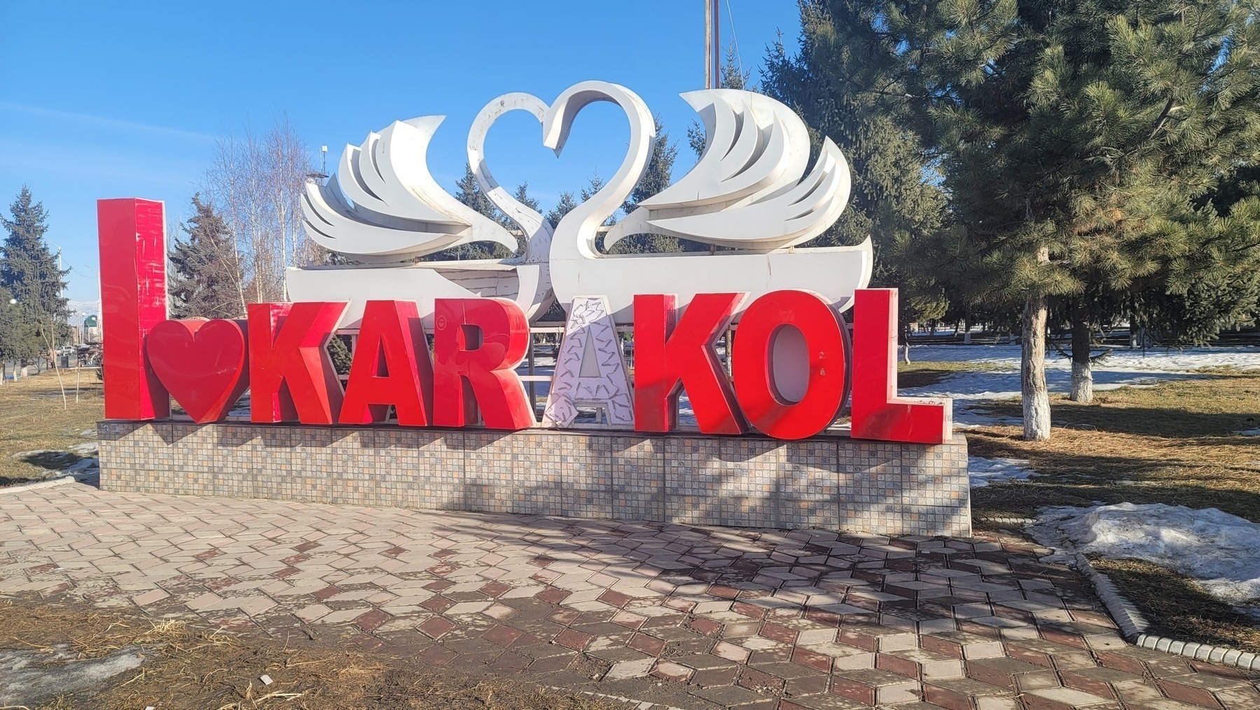sign with big, red block letters that reads 'I (heart) Karakol', but only a thin, white A where the second A should be. above and behind Karakol are two white swans in the same block style as the sign, facing each other so that their heads make a heart