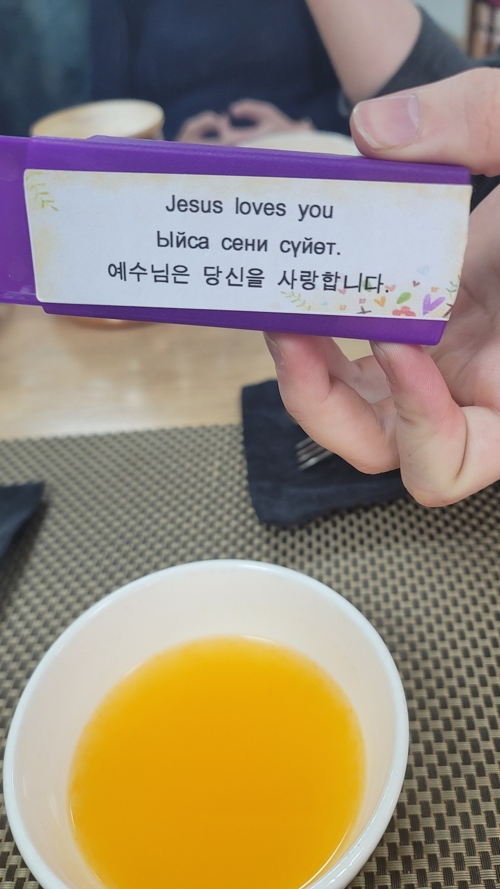 hand holding a purple bandaid case with a white sticker in front that reads "Jesus loves you" in English, Kyrgyz, and Korean