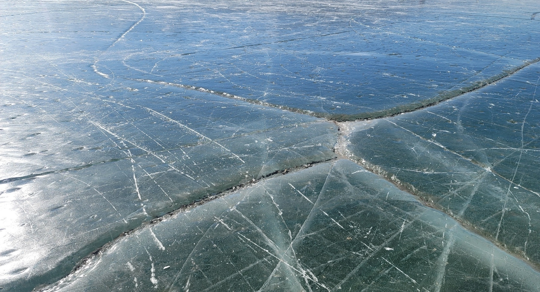 green/gray lake surface with white ice skate cuts and large cracks forming a cross