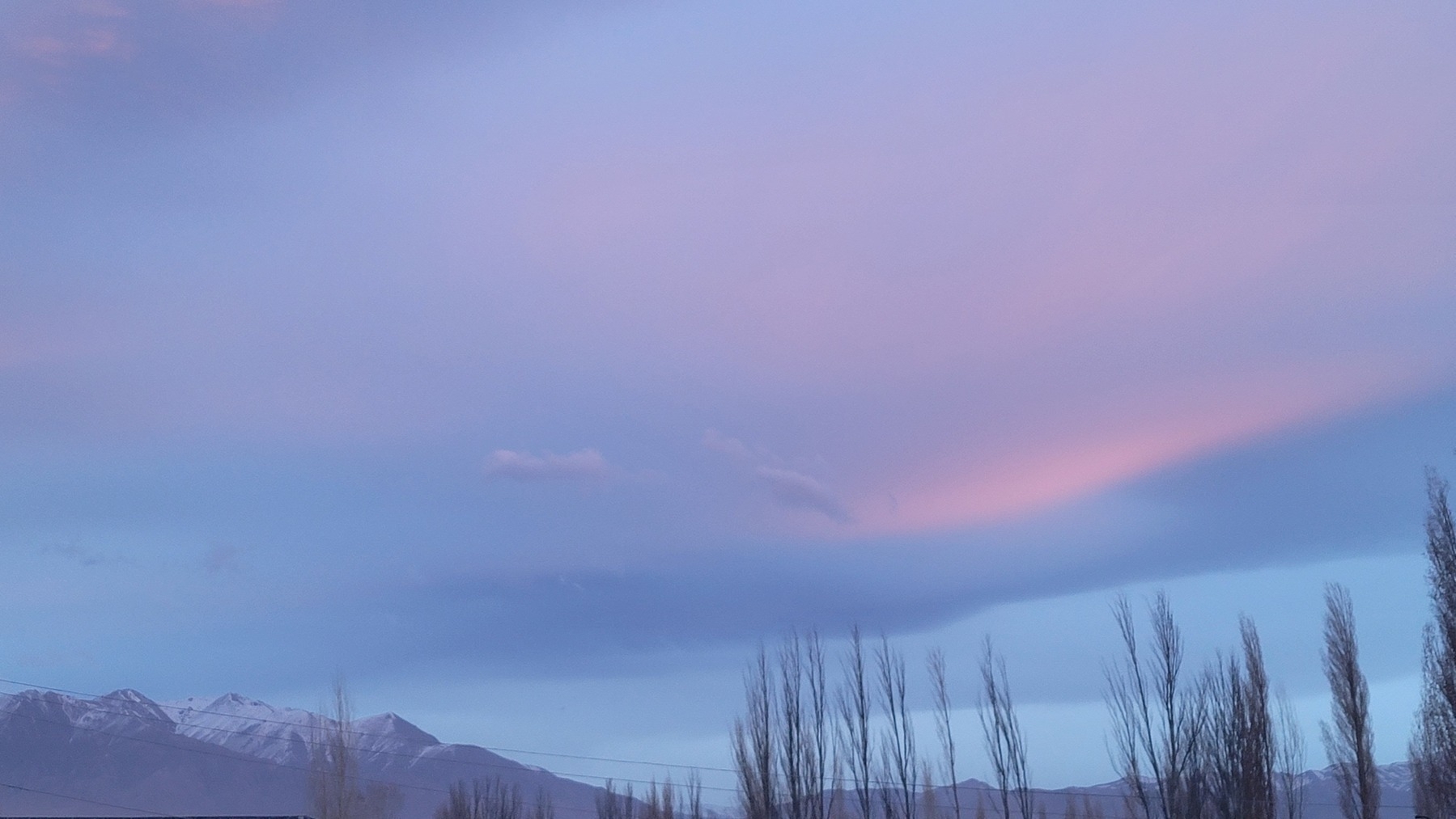sky which looks like 3 layers of blue and a layer of pink over it. mountains in the lower left corner with power lines visible in front of them. trees sticking up from the bottom right
