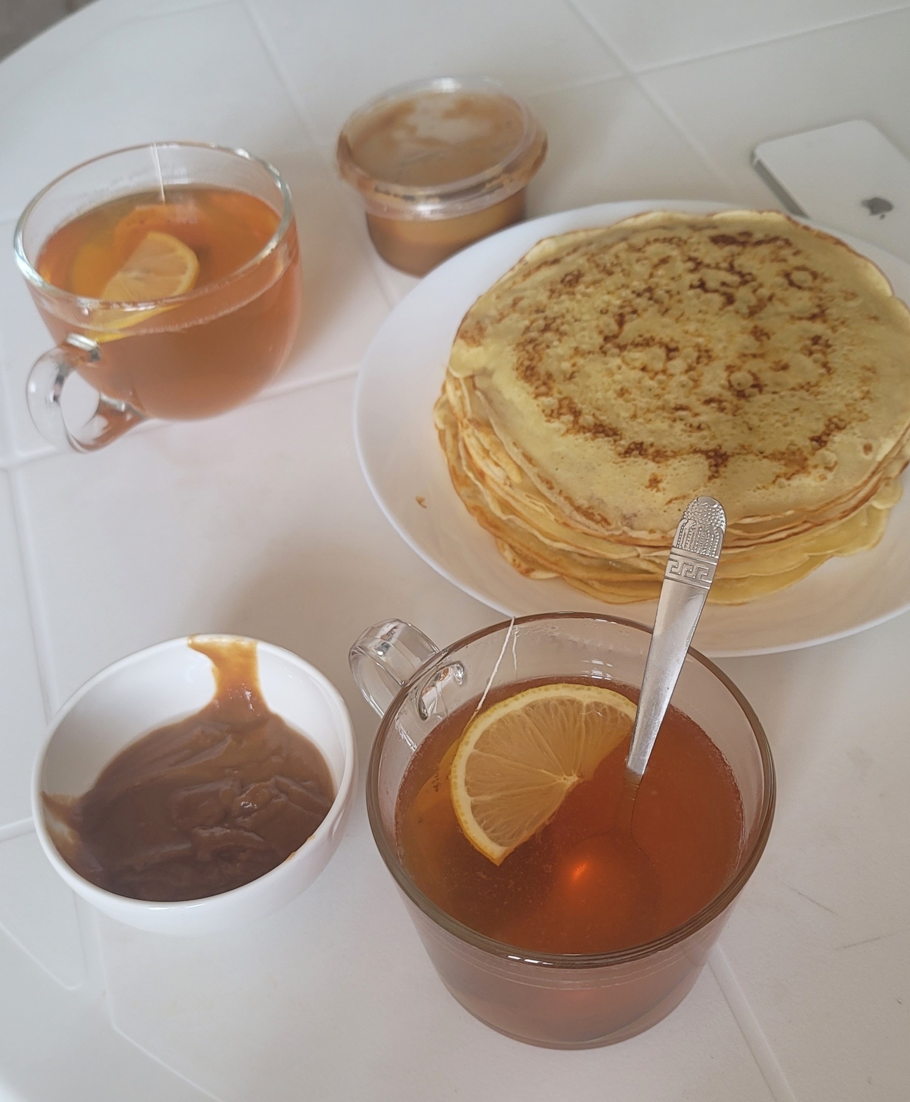 two cups of tea with lemon slices; a plate of blini (Russian pancakes) and a bowl with сүмөлөк, a brown pudding-like dessert (also a plastic container with сүмөлөк)