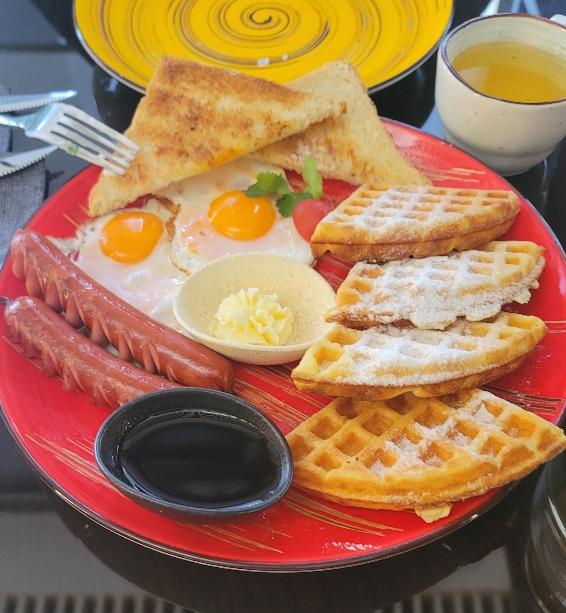 plate with two small pieces of toast, two small fried eggs, a small waffle cut up into 4 pieces, and two sausages. plus small containers of butter and syrup, and a cup of green tea in the top right