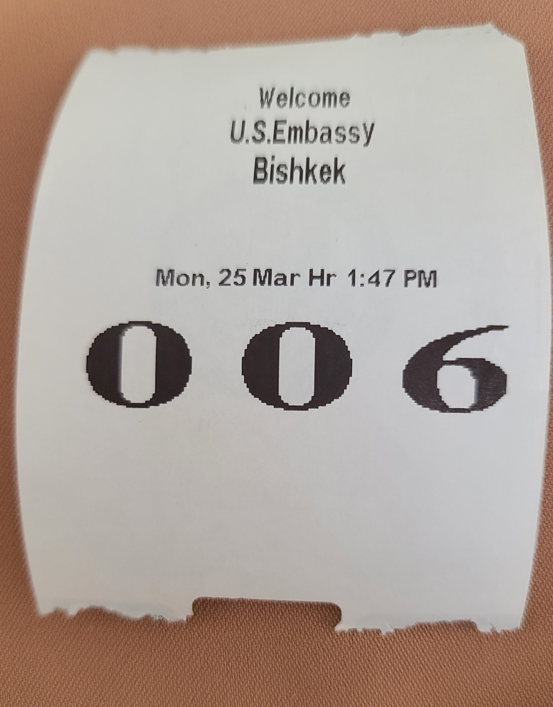 white receipt-looking paper with black text: "Welcome. US Embassy. Bishkek. Mon, 25 Mar Hr 1:47 PM. 006"
