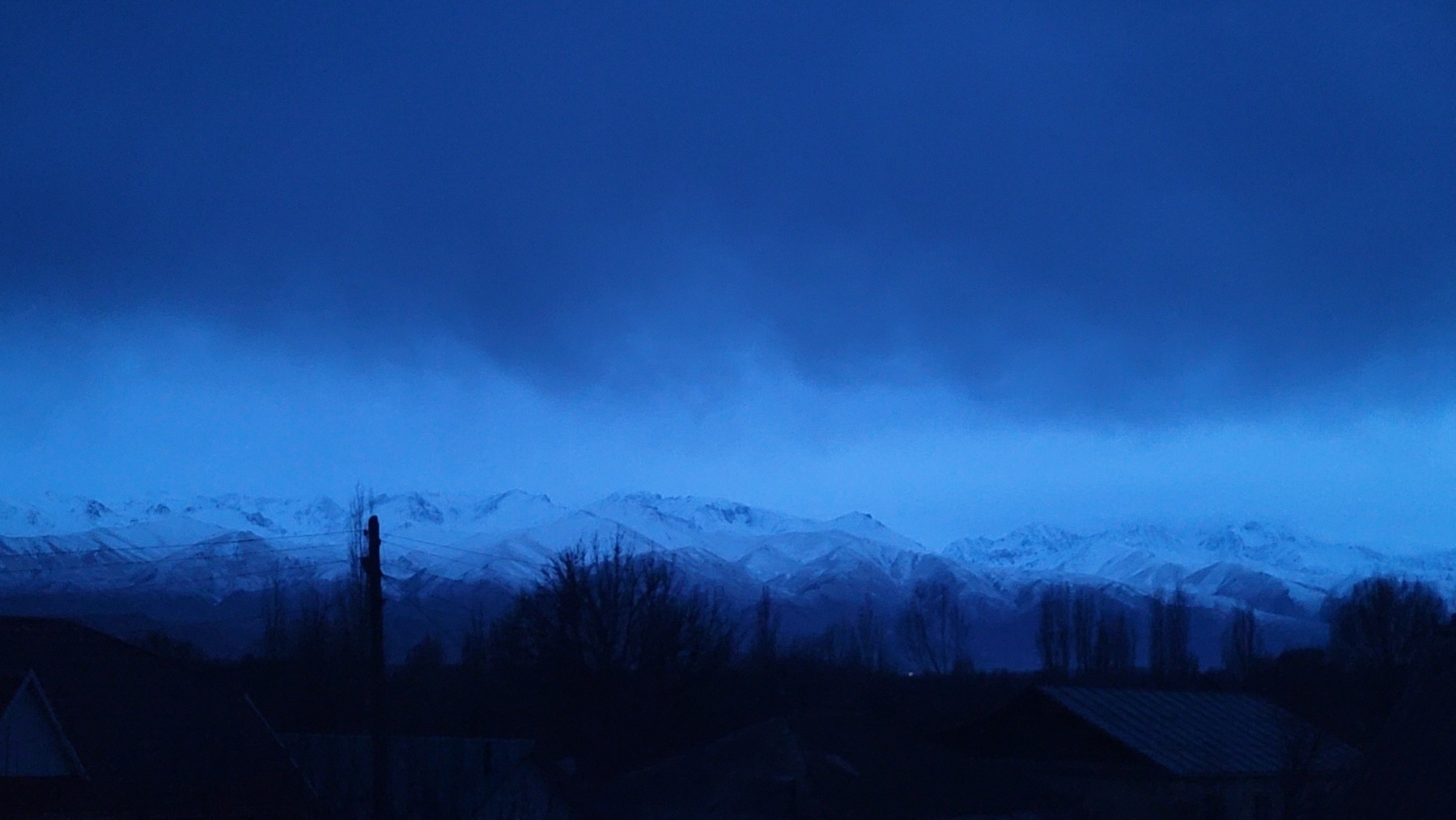 snow-capped mountains against a blue sky after sunset, with dark clouds hanging in the sky almost just above the mountains. silhouettes of trees, houses and a powerline in front of the mountains