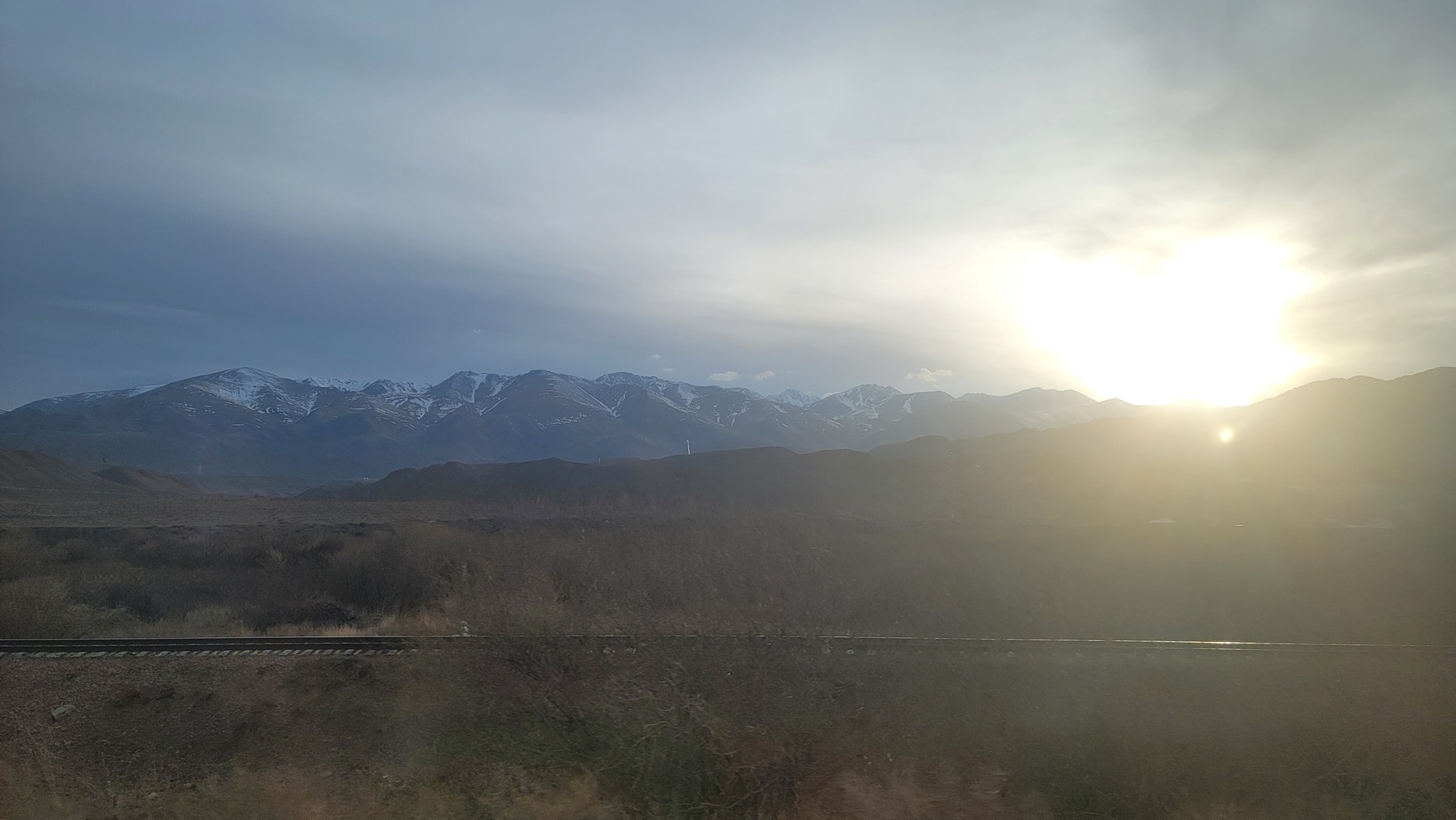 train track running from left to right, with mountains and a shining sun in the background (in the evening)