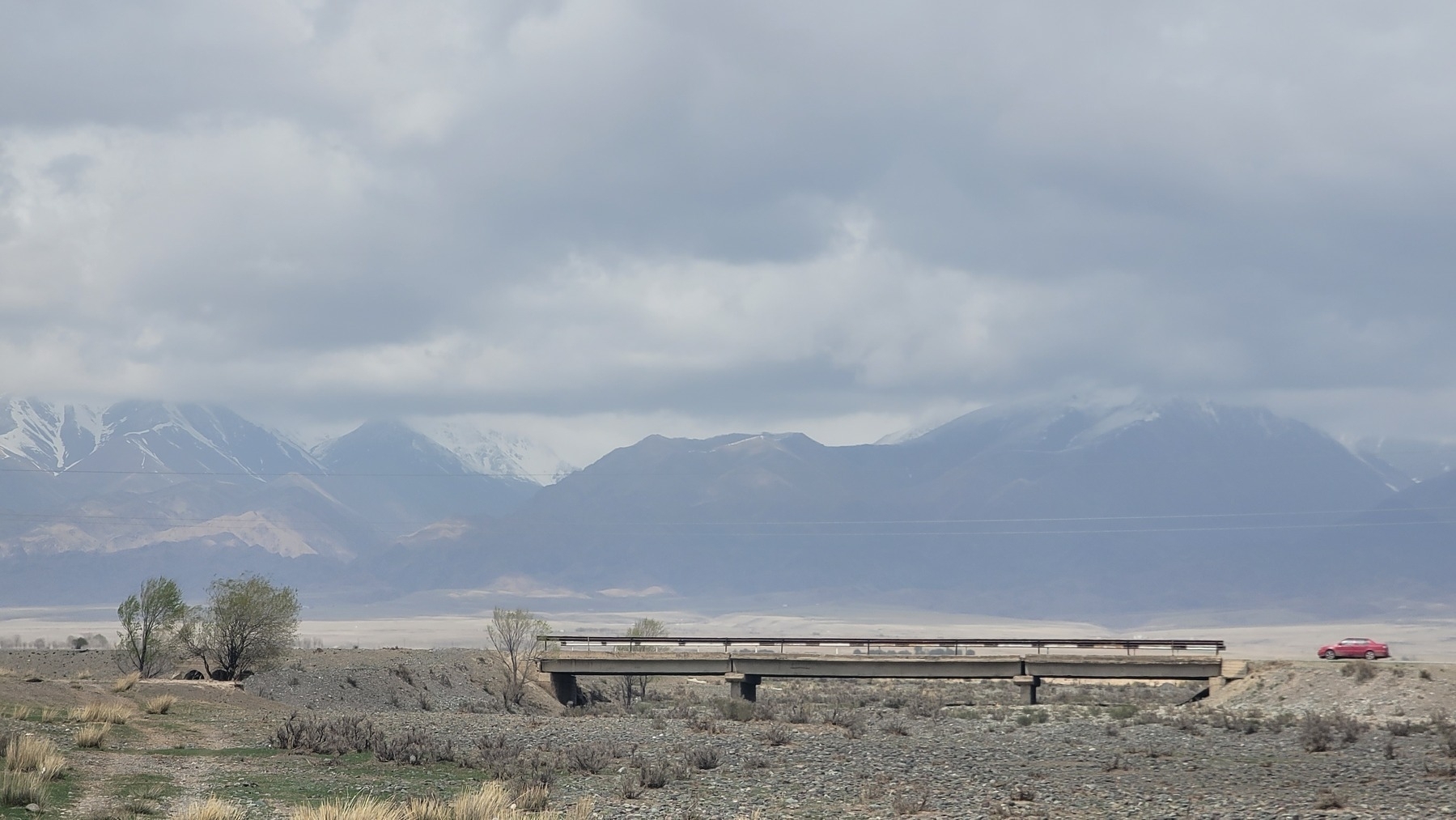 mountains in the background, bridge in the foreground over a dry riverbed. a red car parked to the right, off of but next to the bridge 