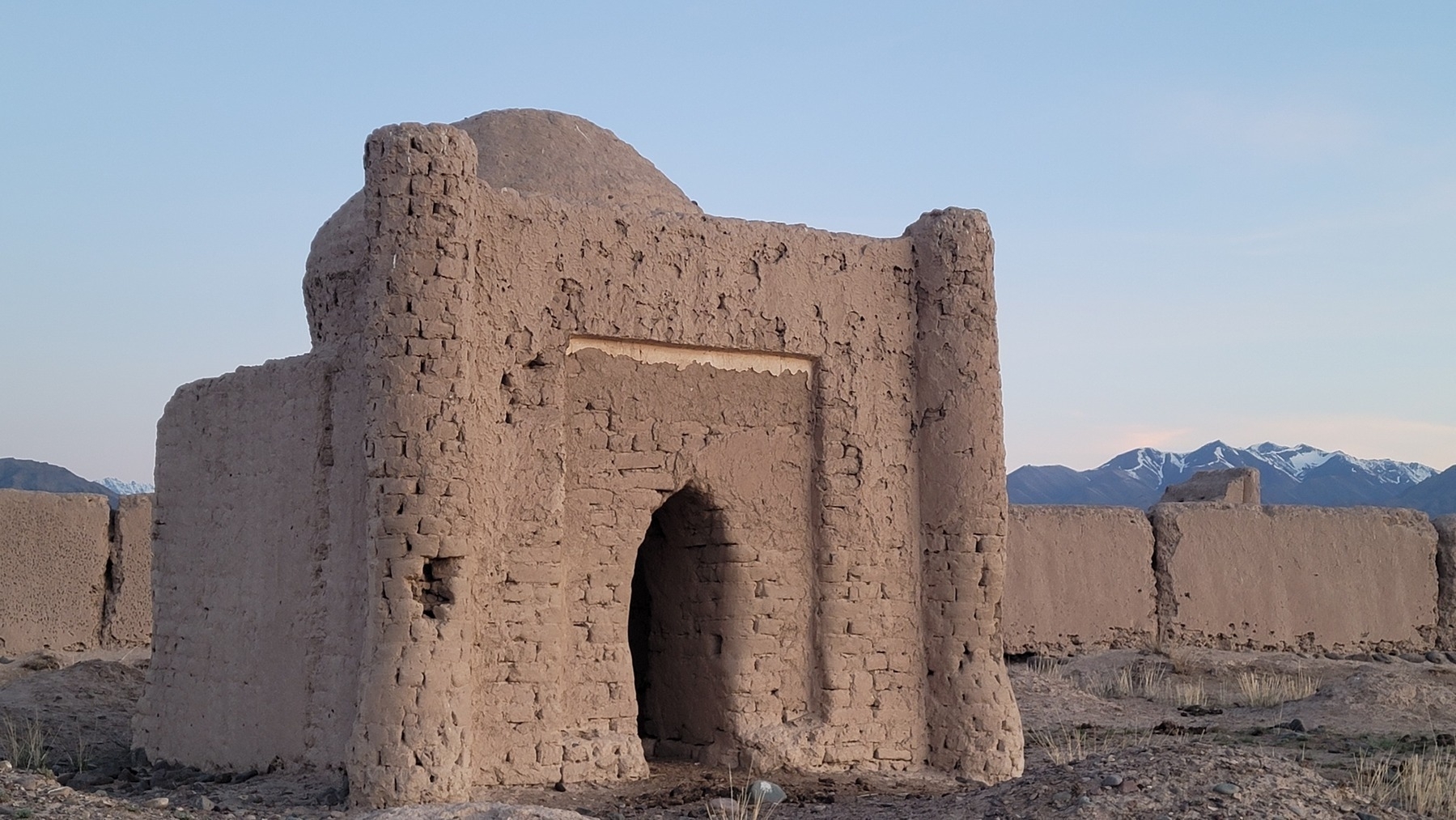 small ancient-looking building made of brown stone/clay(?) with a small domed roof and, a slightly higher wall in front with columns on the right and left sides. wall of a small enclosure made of the same material visible behind, and snow-capped mountains behind that