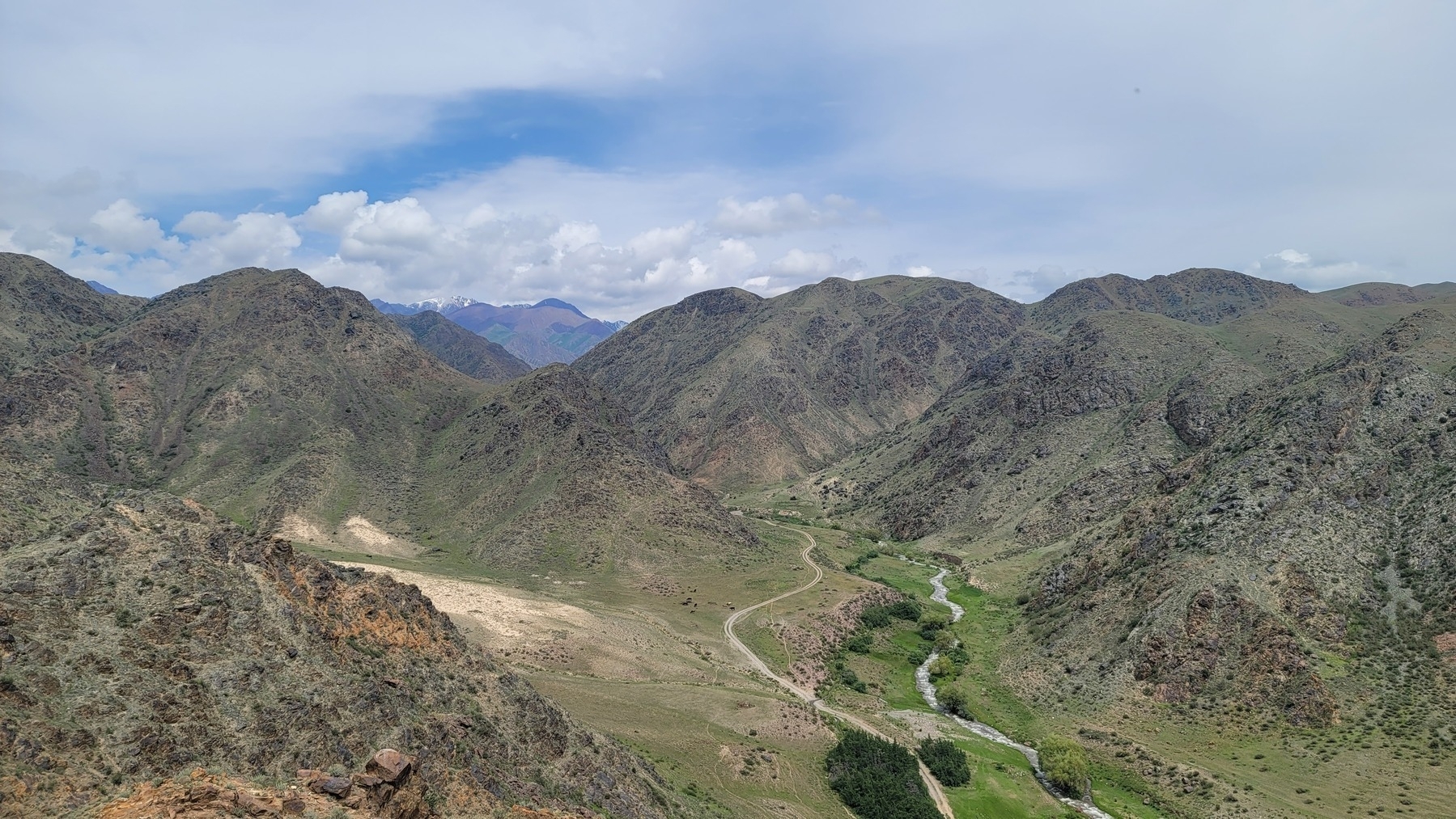 view from the top of a small mountain looking down at a river, dirt car tracks, and other mountains