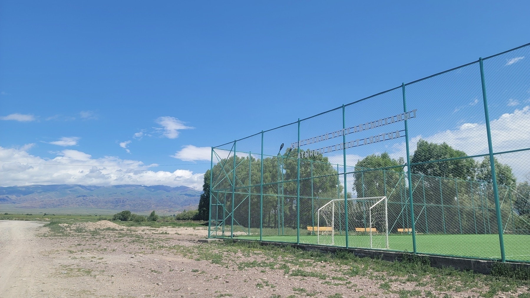 small football (soccer) field with a tall fence all around it, new wooden benches along one side of the field. small mountains visible in the background on the left