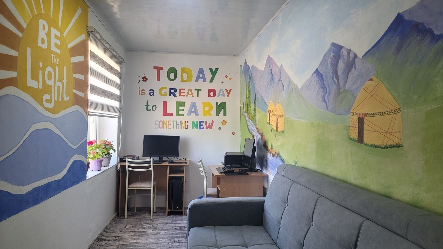 room with two desks with computers, a couch, yurts in the mountains mural on one wall, "today is a great day to learn something new" in colorful letters on one wall, and "be the light" sunrise over mountains mural on another wall