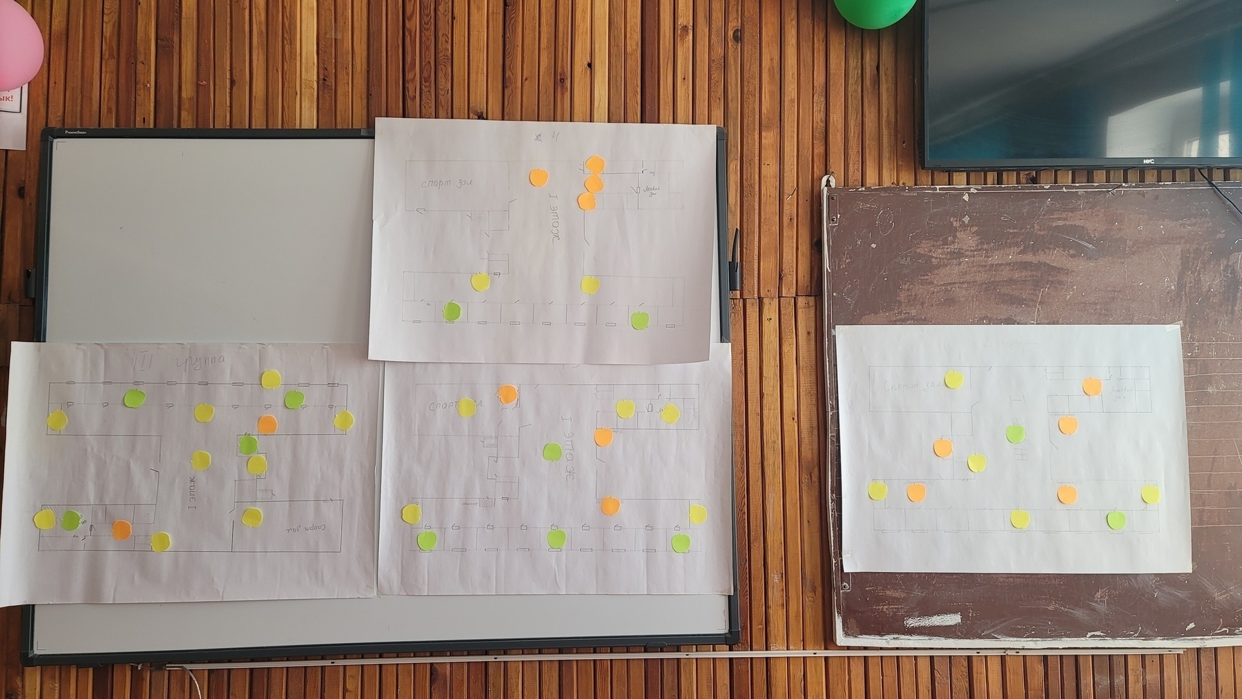 smartboard and brown chalkboard hanging on a wooden wall, with 4 school map paper posters (with green, yellow and orange sticky notes) taped on top