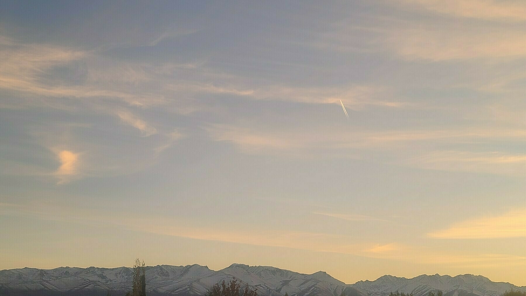 snow-capped mountains below a light blue/yellow sky with a plane and some jet stream behind it