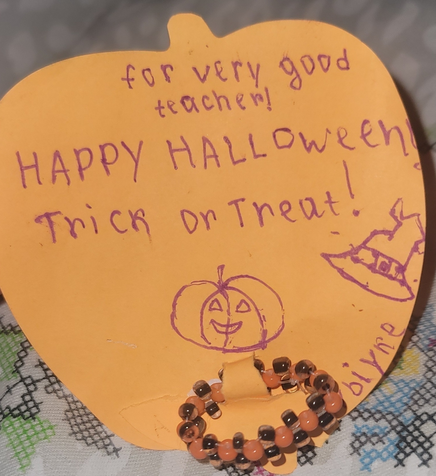 orange, apple-shaped sticky note with a handwritten note reading, 'for very good teacher! Happy Halloween! Trick or treat!" and a small, orange and black bead ring attached