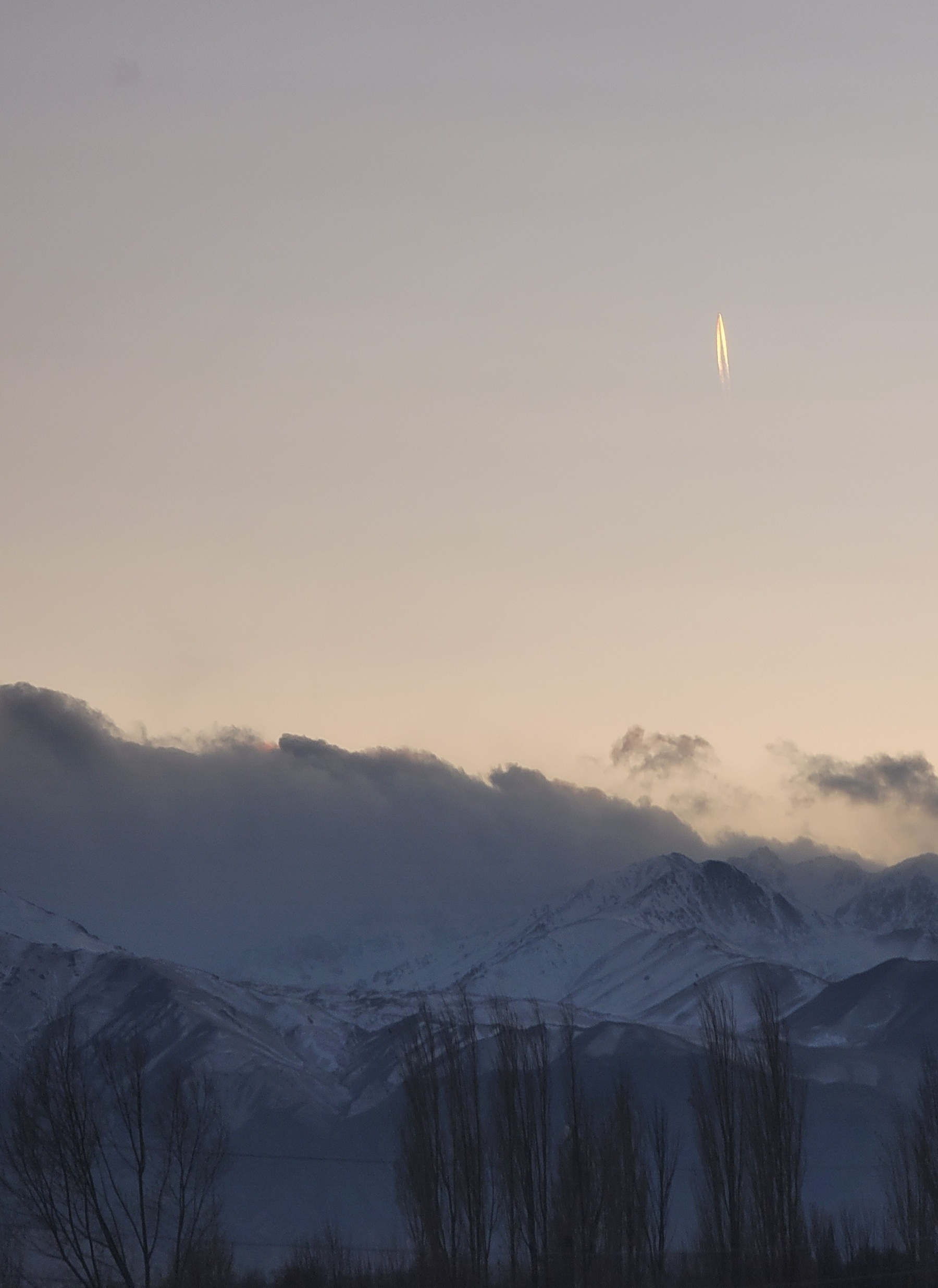 snow-capped mountains with dark clouds above; light orange sky with a plane going straight up, jet stream coming from it. around sunset