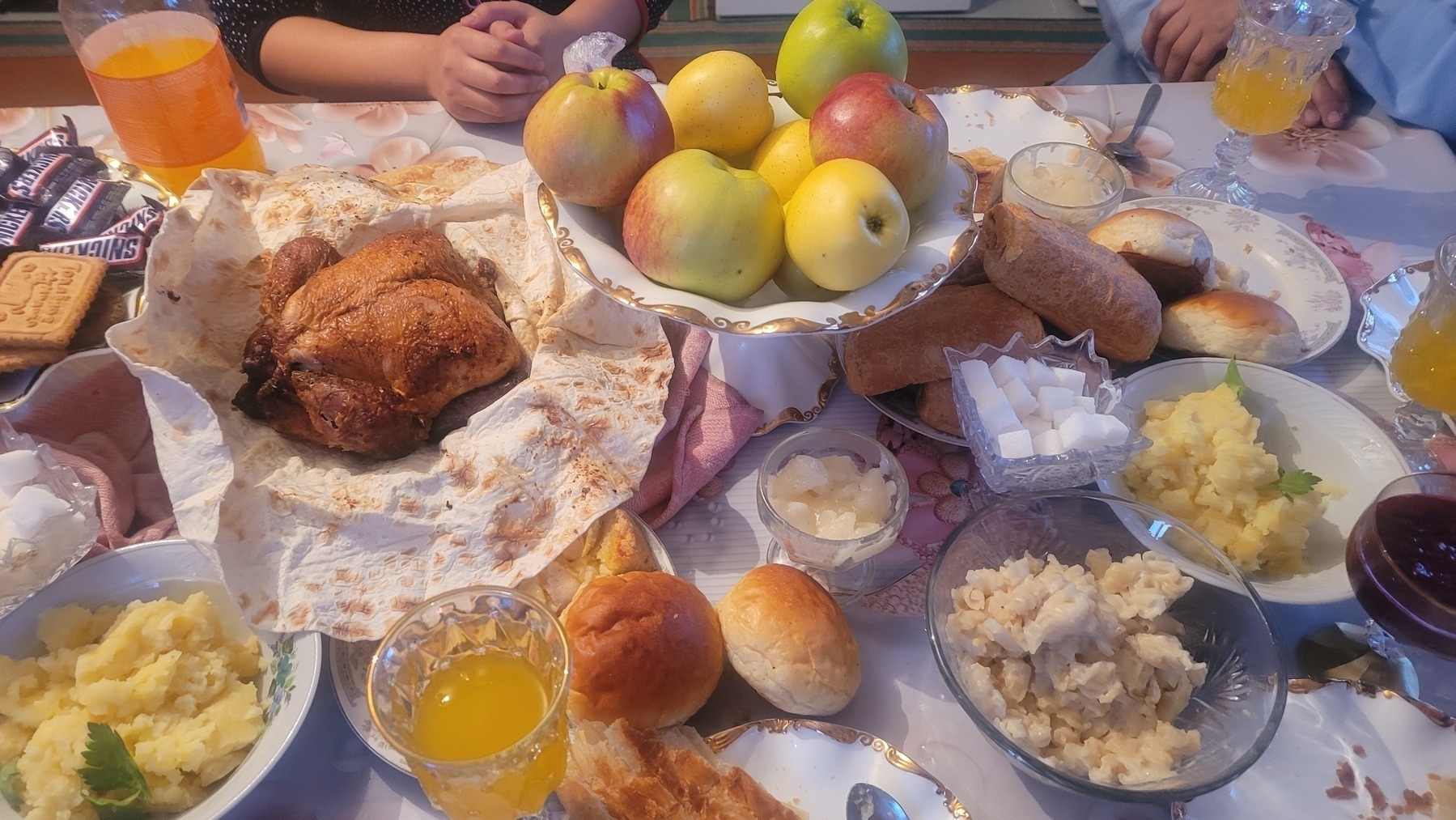 tabletop with food and drinks including mashed potatoes, a whole chicken on a slice of lavash, macaroni and cheese with white cheese, dinner rolls, Kyrgyz kattama flatbread, apples, Samsa, mini snickers