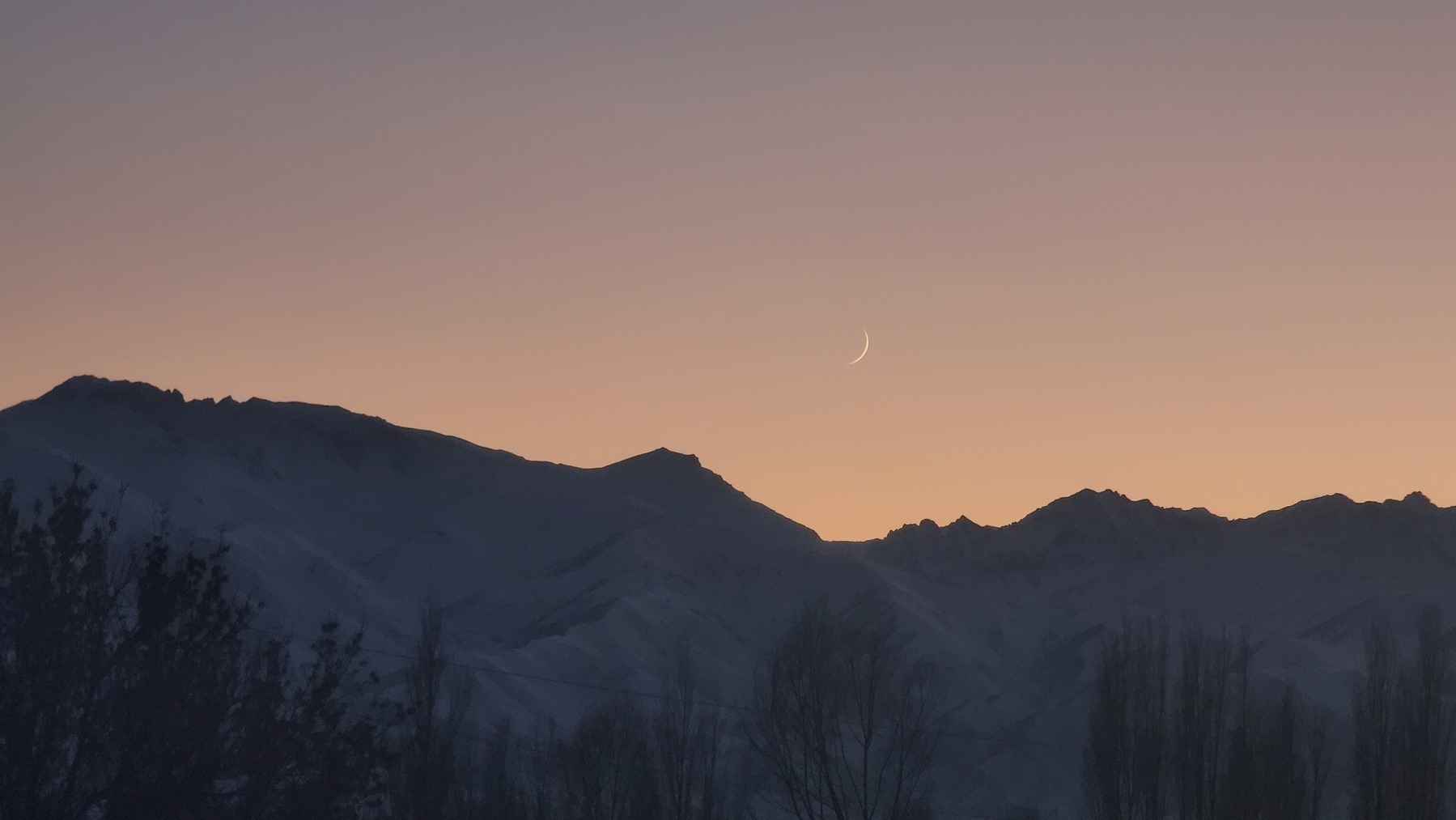 tiniest sliver of a waxing crescent moon in a pale pink/orange sunset sky, with mountains below