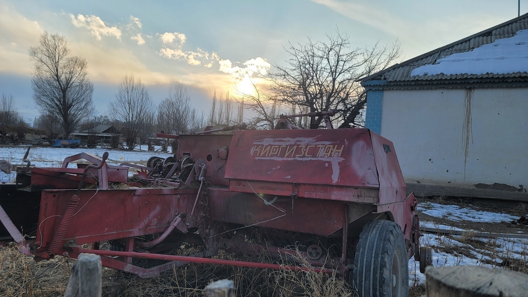 red farming machine with "Kyrgyzstan" written on it in Cyrillic letters, next to a house