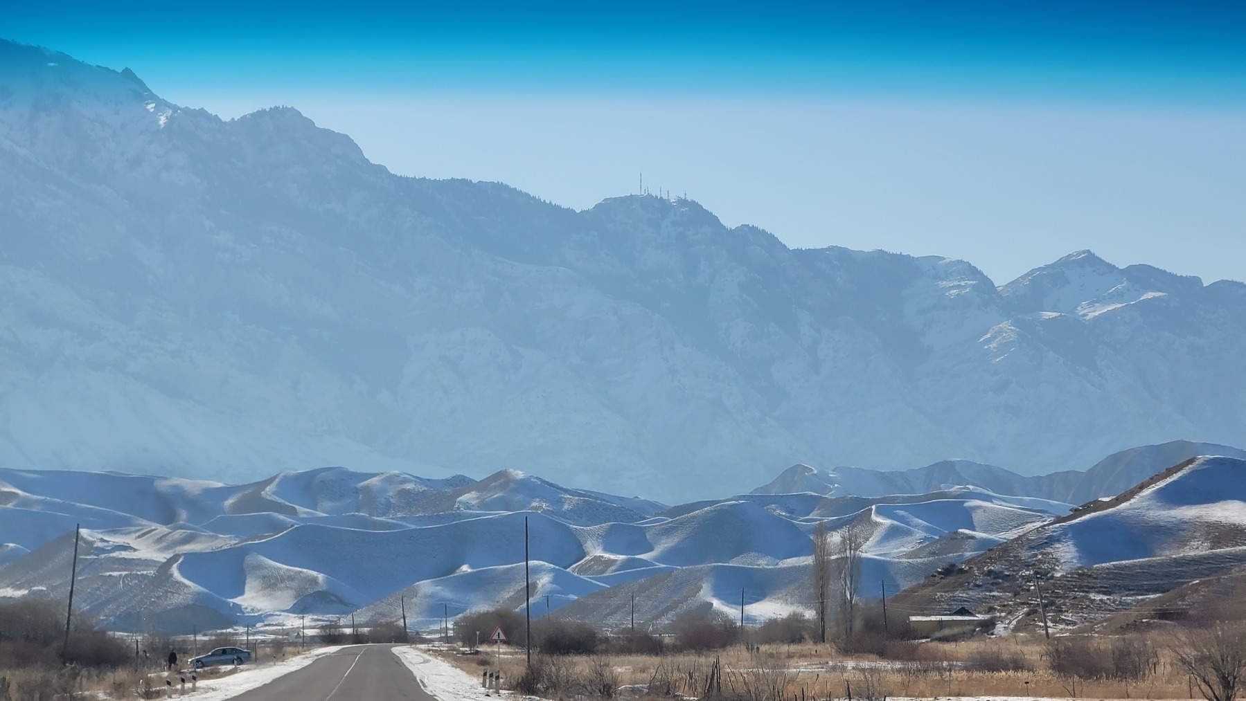 road snaking right next to a series of small snow covered hills, with mountains behind in the distance 