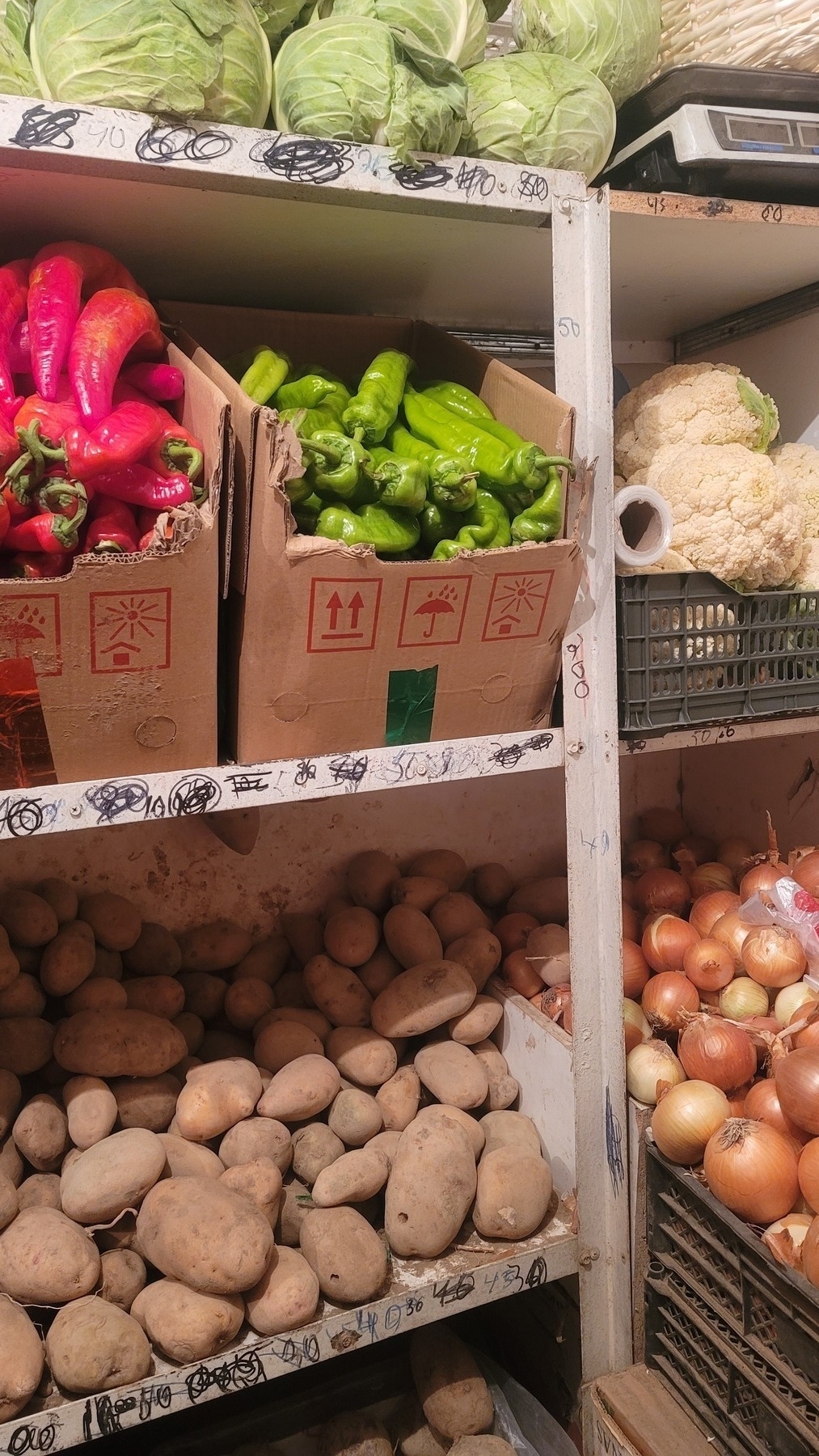 vegetables on white shelves that have many scratched out numbers (prices) on them