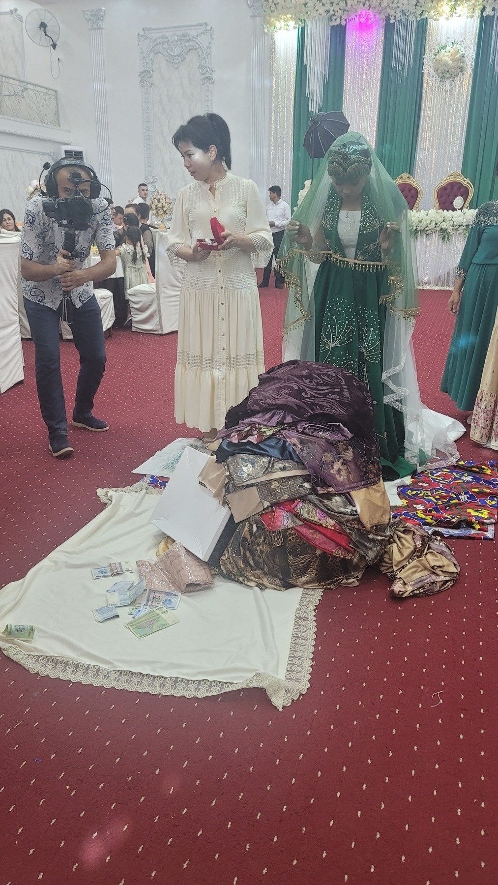 bride with a green coverup over her white dress and a light green veil standing in front of a pile of scarves and other gifts