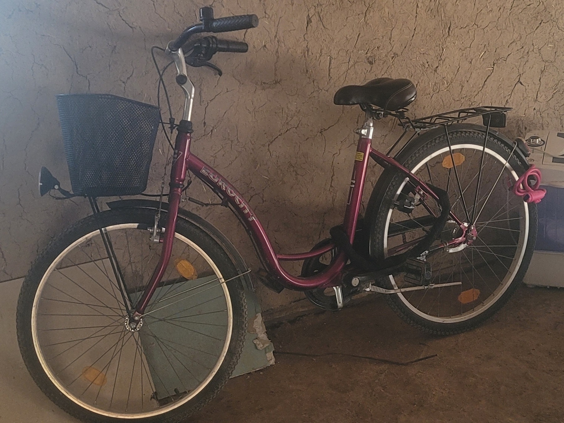 maroon bike with a black basket standing against a wall. two bike locks (one pink, one black) on it