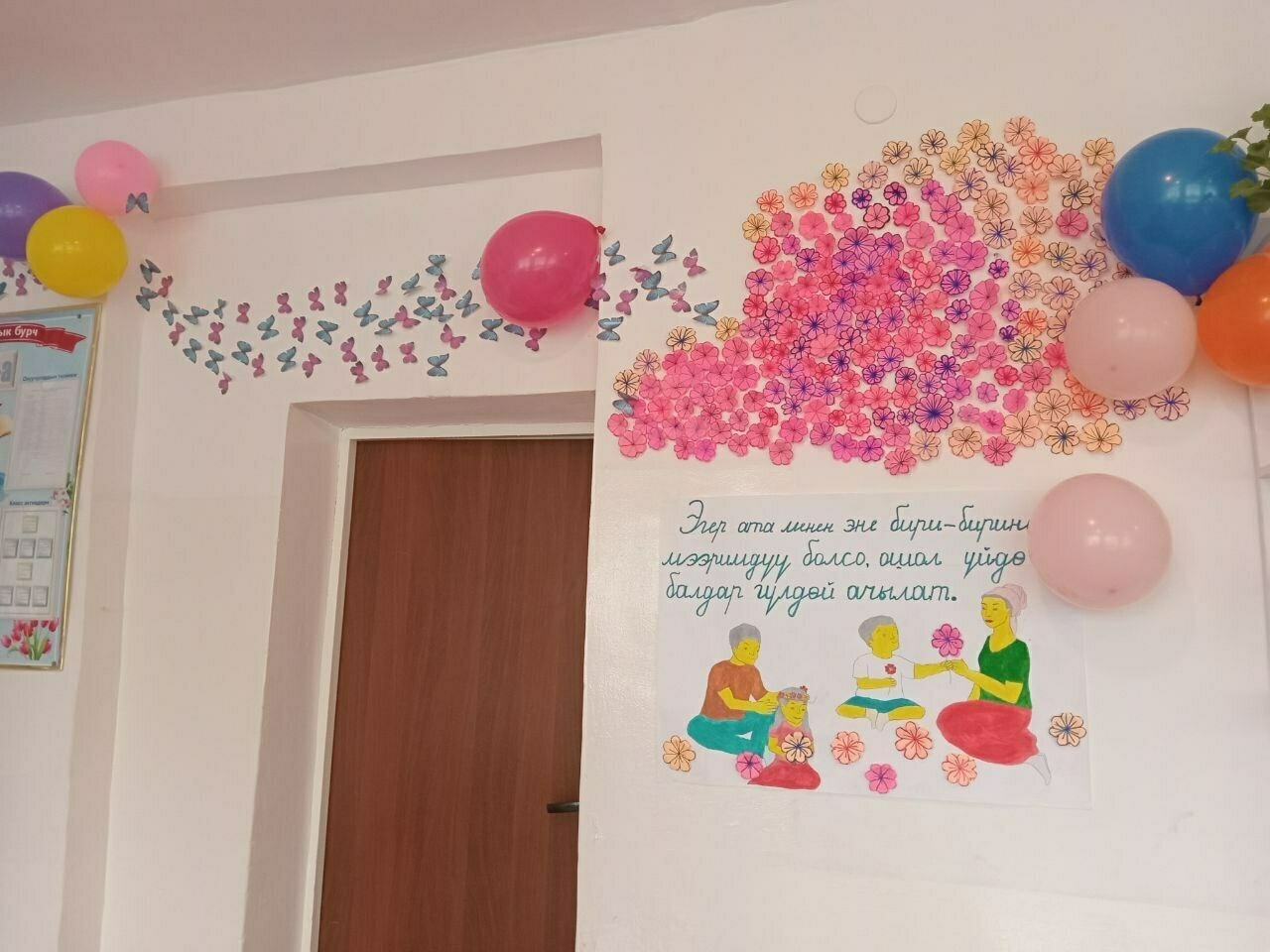 trail of colorful paper butterflies and flowers on a classroom wall above the door; colorful balloons hanging on the wall and a poster painted with a picture of a family and some Kyrgyz words on it also on the wall 