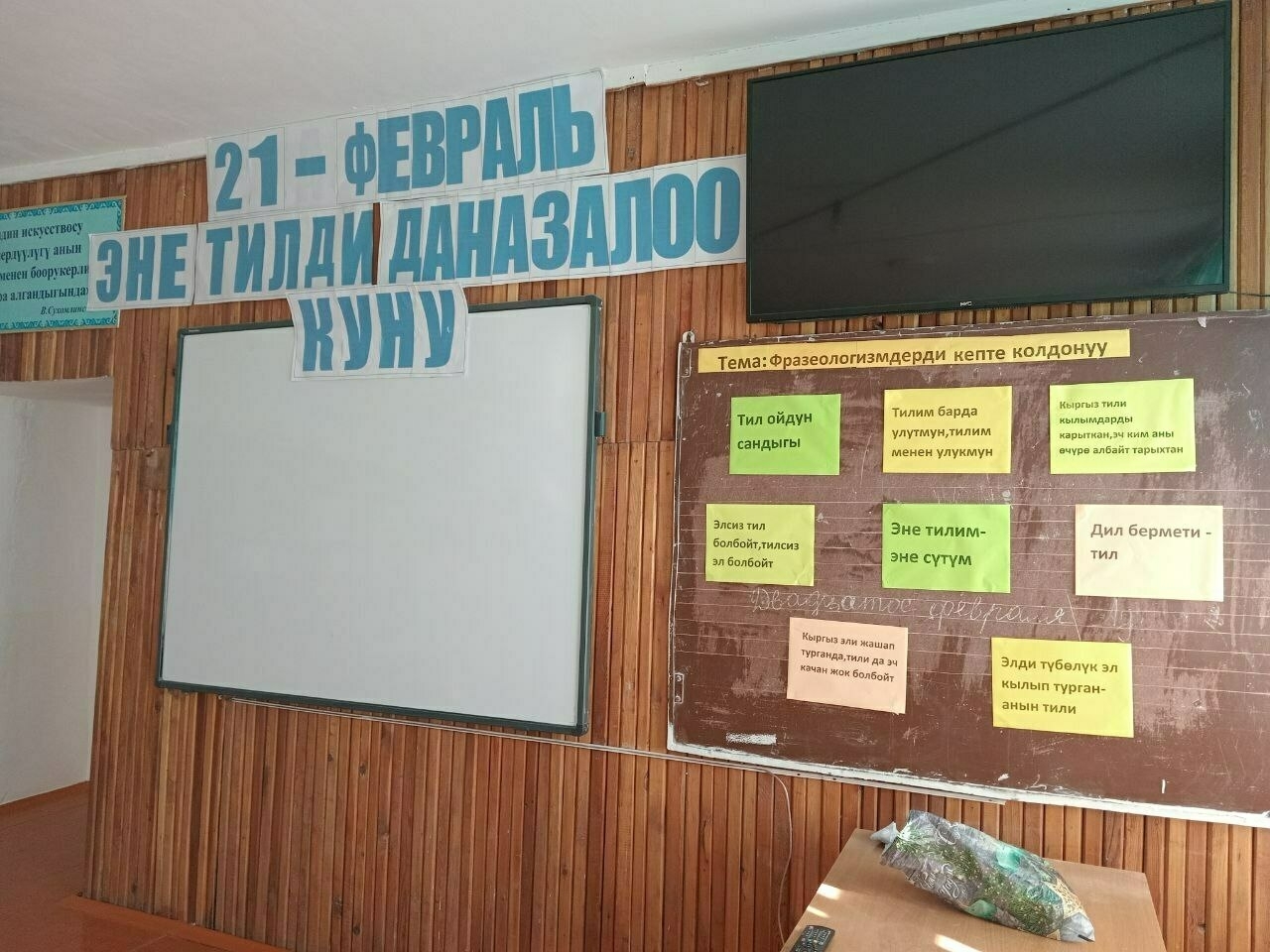 classroom with "21st February Mother Language Day" letters taped the wall and top of a smartboard; to the right a TV on the wall; below the TV an old brown chalkboard with colored papers taped to it with quotes in Kyrgyz 