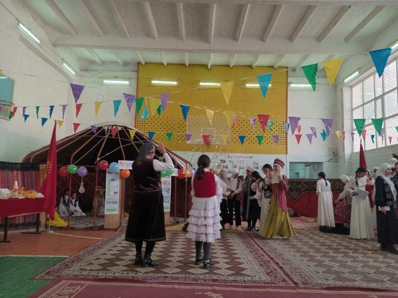 women in traditional Kyrgyz clothes on rugs in a gym. also a yurt structure (without the felt) set up