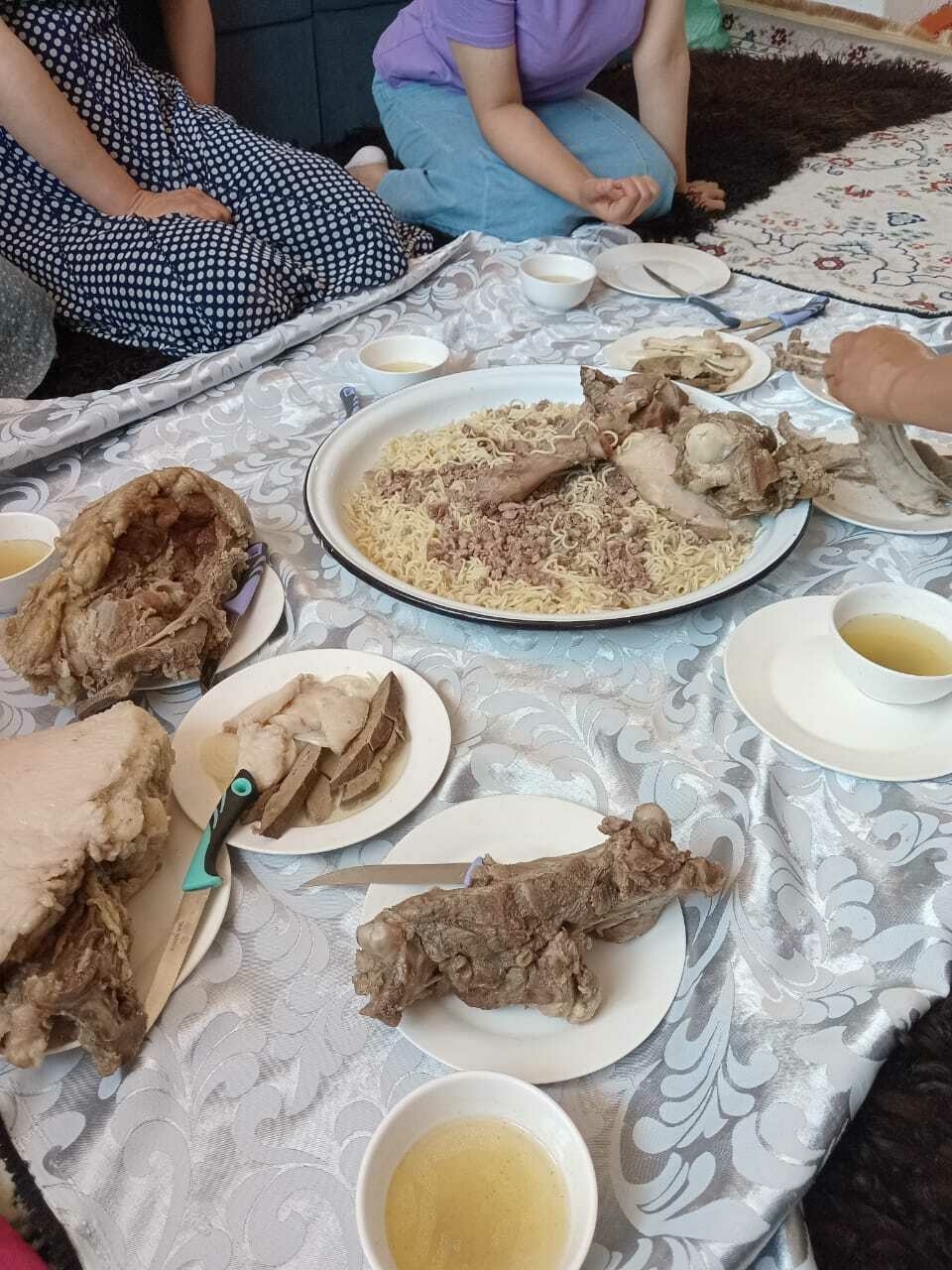 plates of meat and fat spread around a table mat on a floor, a big plate of meat and noodles (beshbarmak) in the middle, and tea cups filled with broth spread around the table mat