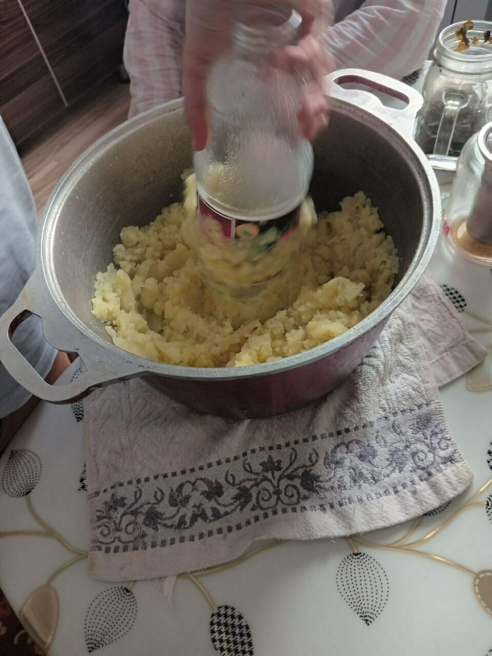 person standing and holding a glass bottle to mash potatoes in a red pot