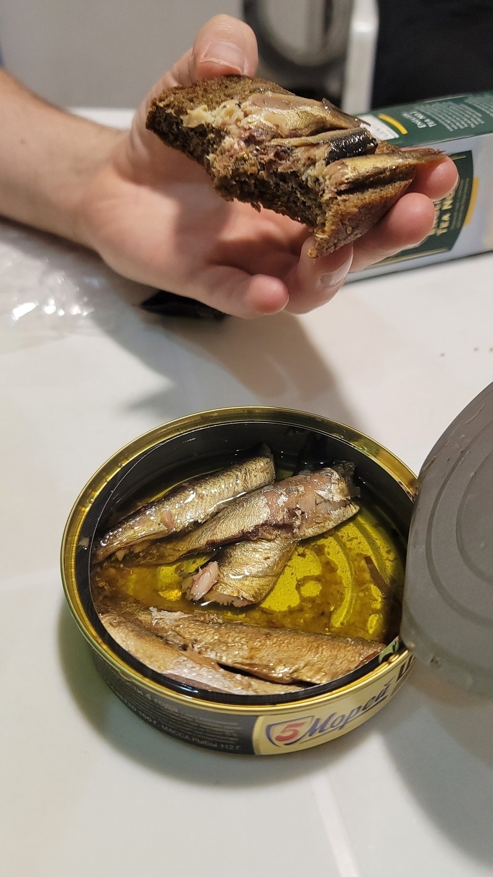 can of sprats and a person's hand showing a half-eaten piece of bread with sprats on it