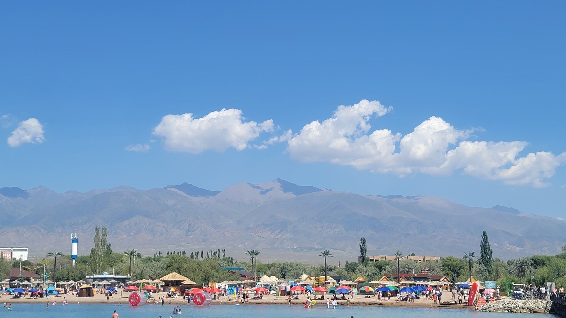 People and umbrellas along a lake beach. Mountains in the background