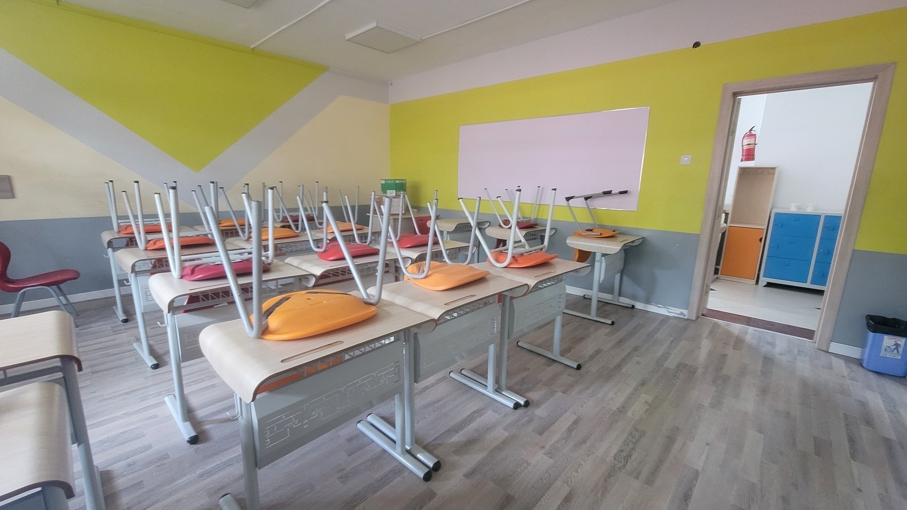 new-looking and bright classroom with yellow/gray walls, light gray floor, and individual desks, each with a red or orange chair