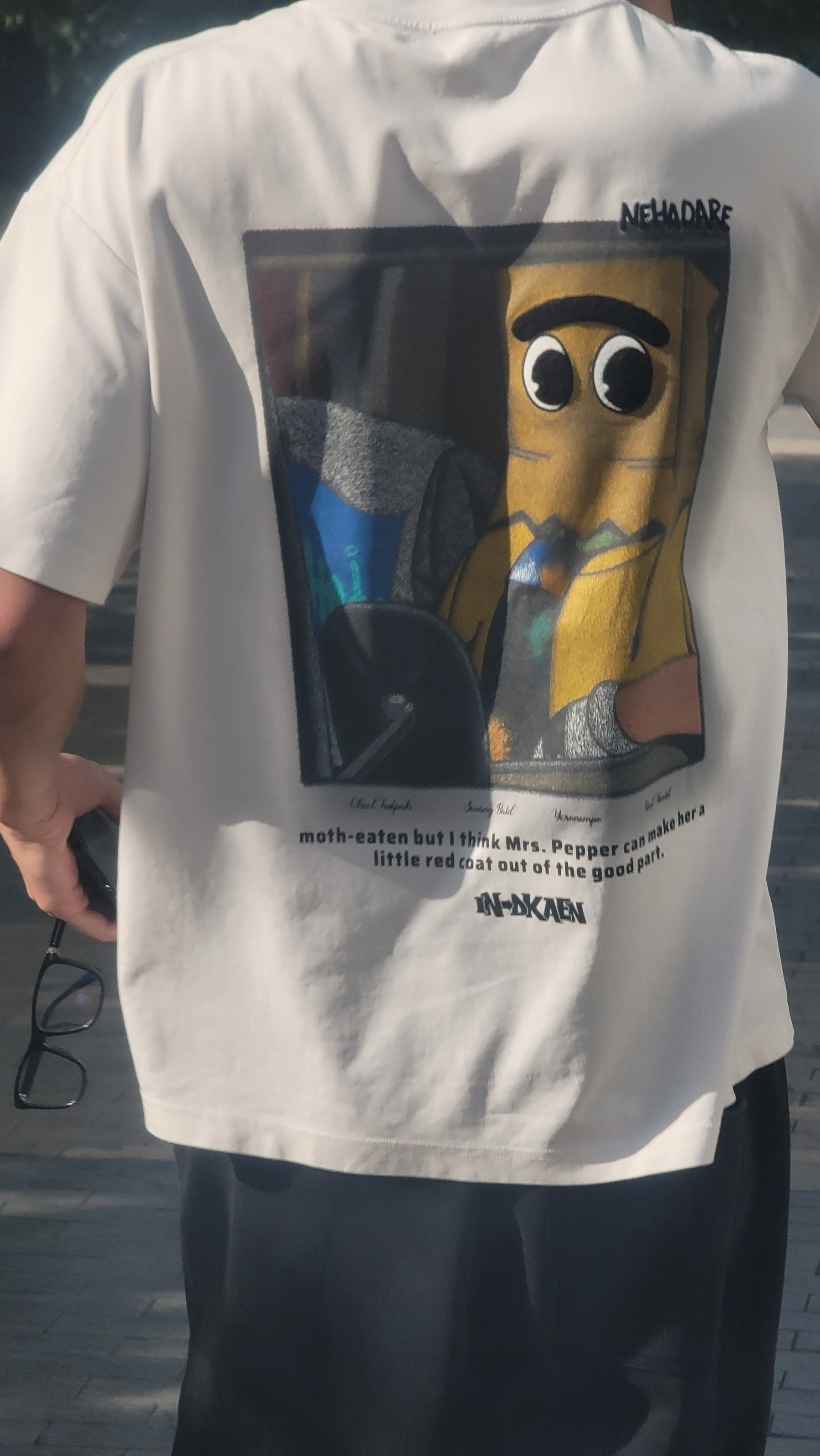 Strange back of a shirt with a picture of a cartoon driver with a unibrow who looks like he is wearing a paper bag over his face, with the text underneath: "moth-eaten but I think Mrs. Pepper can make her a little red coat out of the good part"