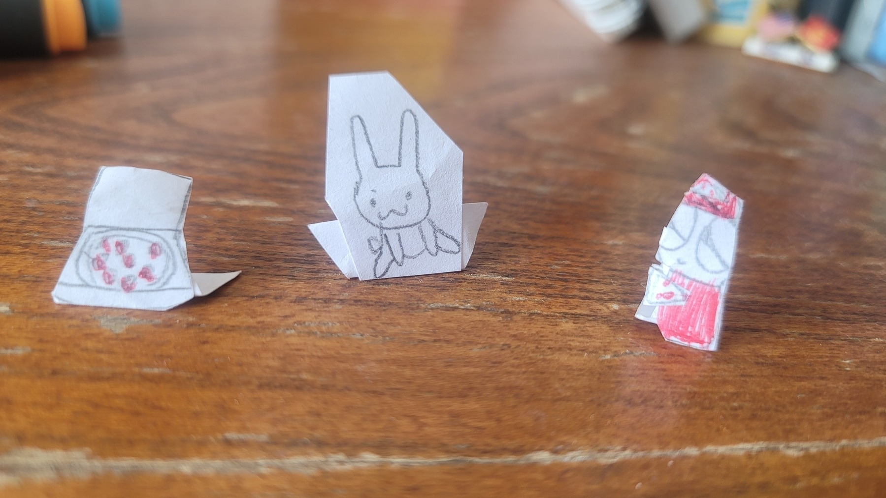 Three small pieces of paper, one with a pizza, one with a bunny, and one with a strange cat-like creature drawn on them. Also folded so that they stand up by themselves on a surface