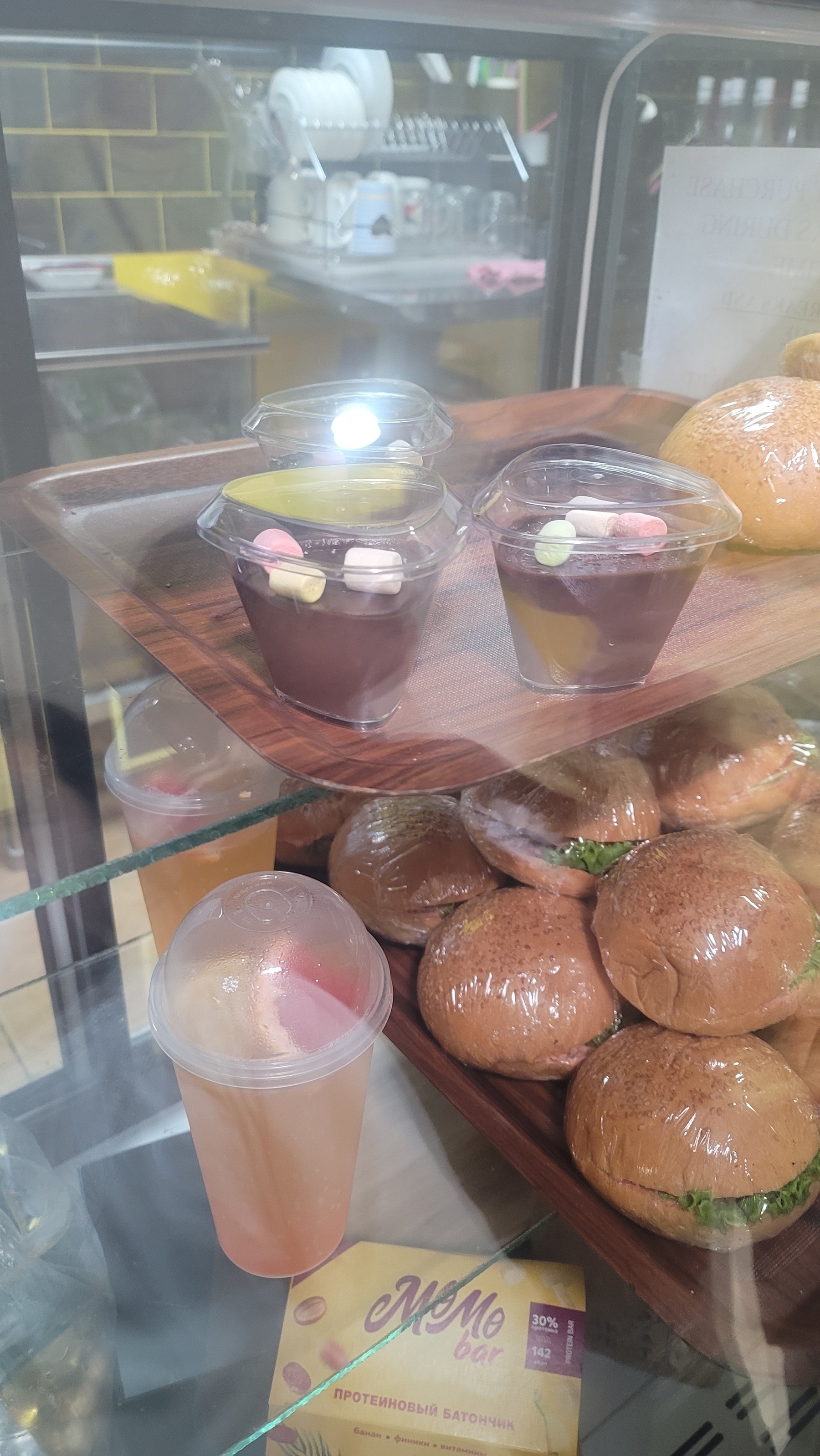 chocolate parfaits with marshmallows on top inside a clear coffee shop display case. Thick, round sandwiches wrapped in plastic wrap underneath