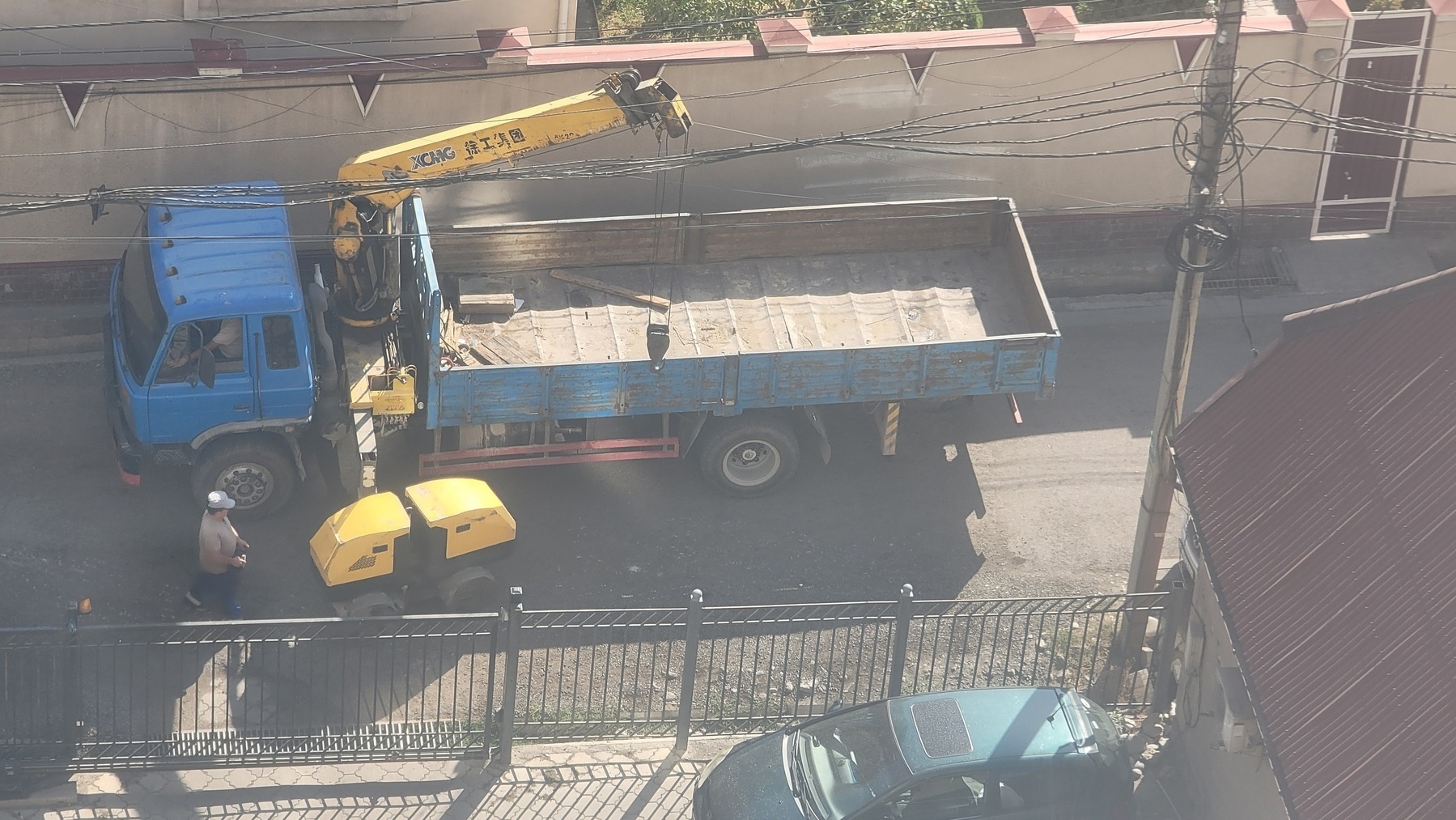 blue truck with a long truck bed. a very small, two-part yellow vehicle next to it, and a man behind the yellow vehicle holding something in his hands and looking at the yellow vehicle