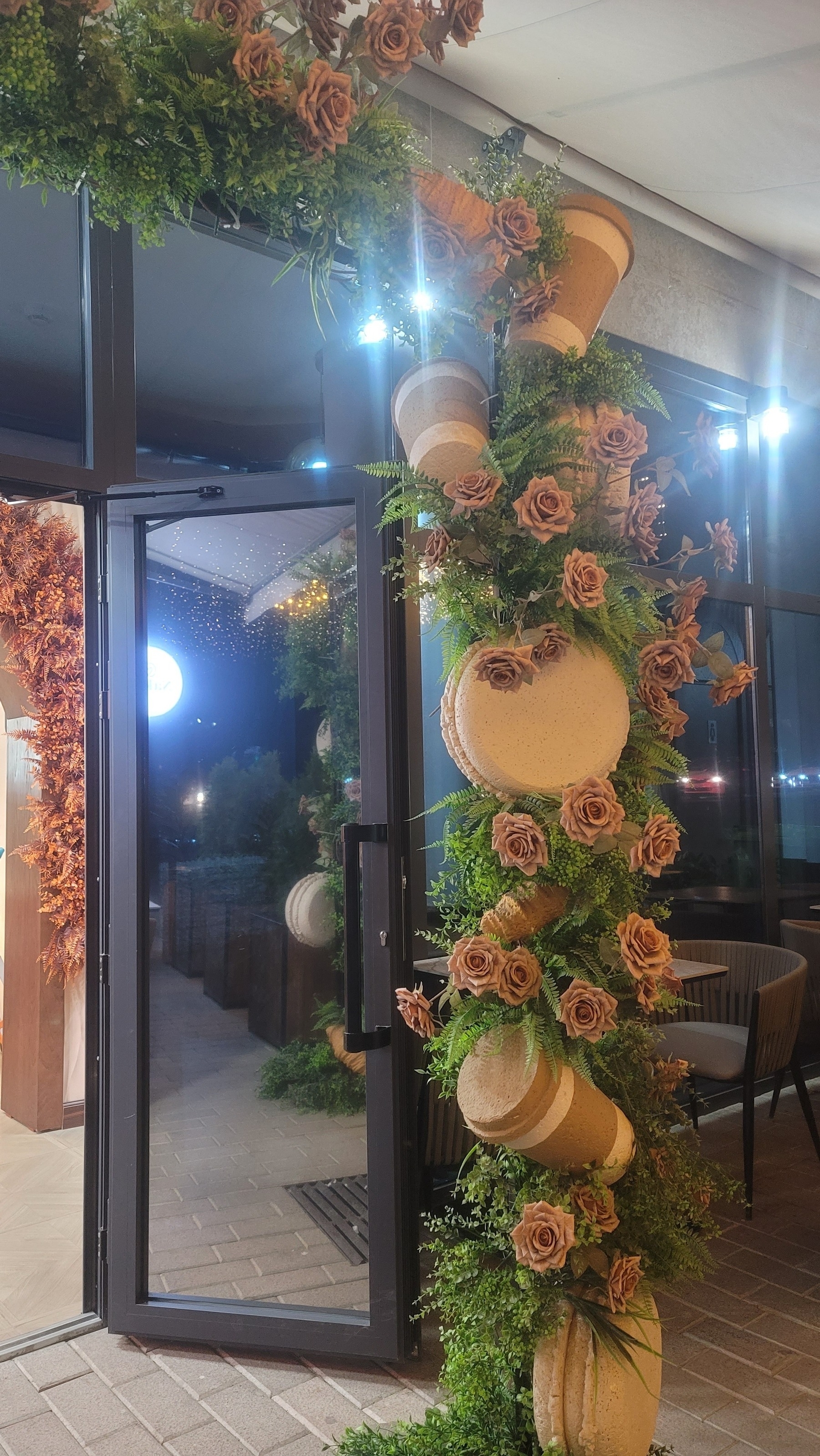 Half of a fake green garland arch in front of an entrance door. Fake flowers, fake macarons, and fake coffee cups within the garland
