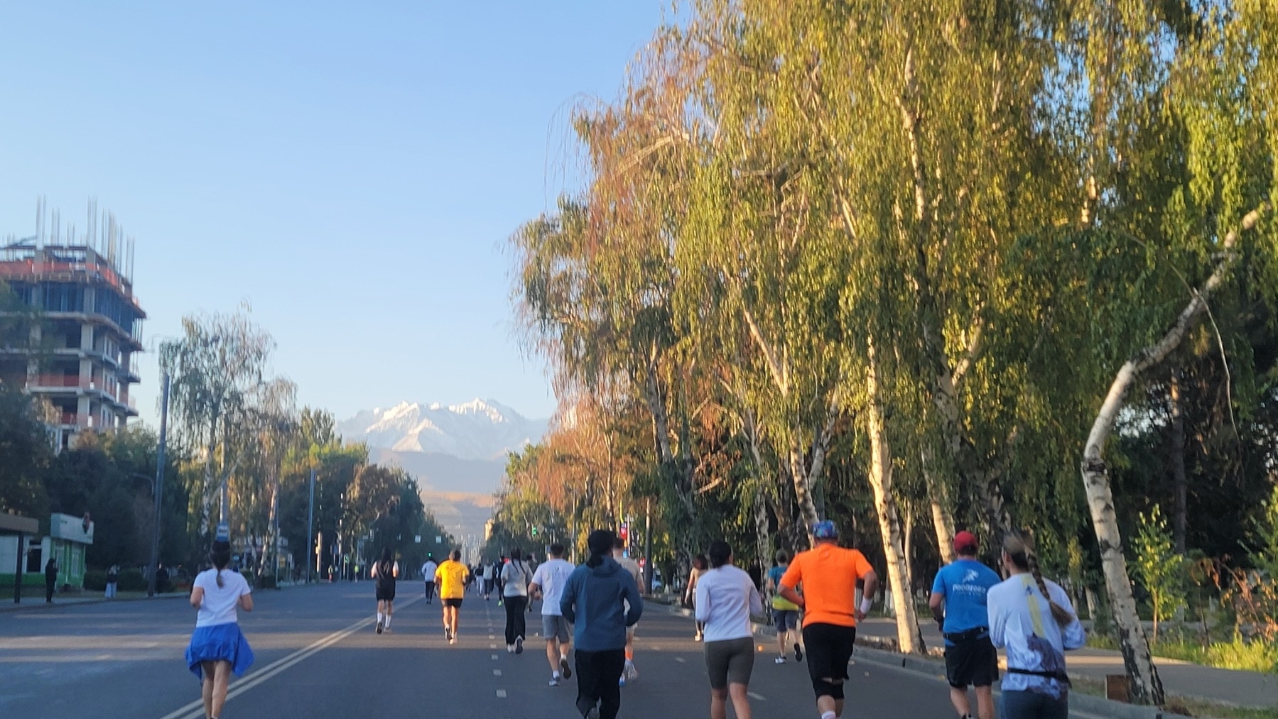 runners on a road. building construction on the left, trees on the right, mountains in the distance down the road