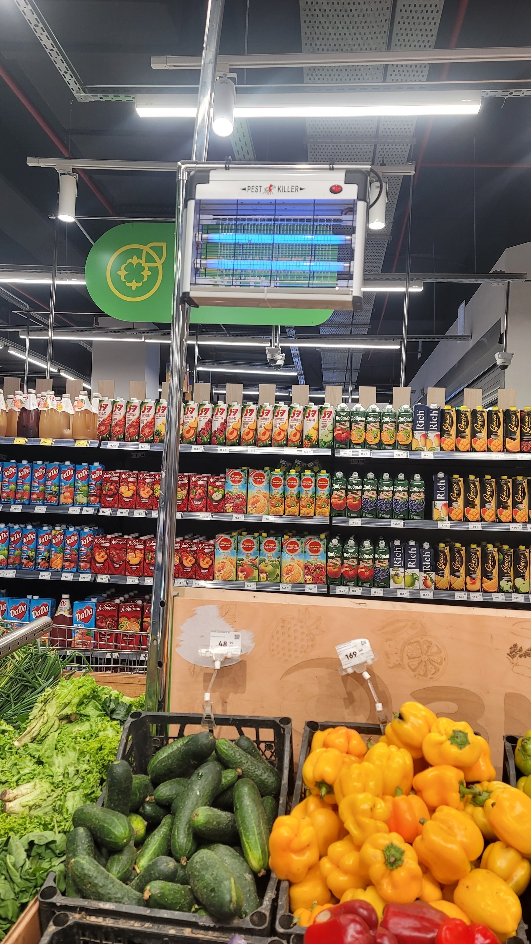 Small bug zapper machine above crates of produce (greens, cucumbers, and yellow/red bell peppers) at a supermarket