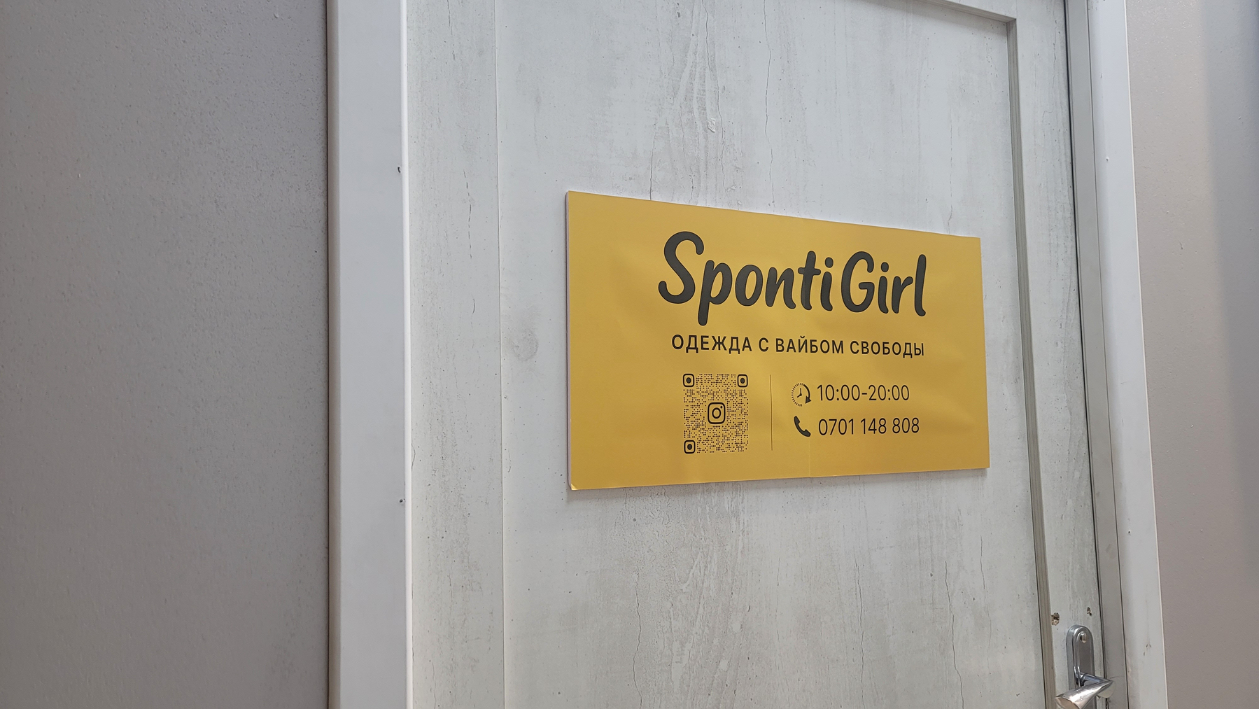 Golden sign on a light gray door that says "SpontiGirl, clothes with free vibes" (the last part in Russian), an Instagram QR code, hours, and a phone number 