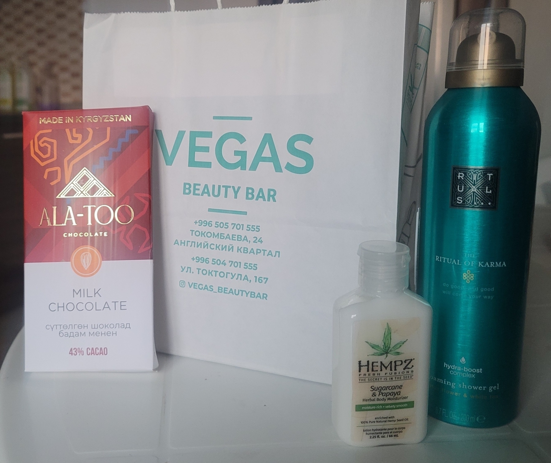 Ala-Too brand Kyrgyz chocolate bar, a white paper bag with the store name Vegas Beauty Bar written on it in teal, a small plastic container of hand moisturizer, and a tall, teal can of shower gel