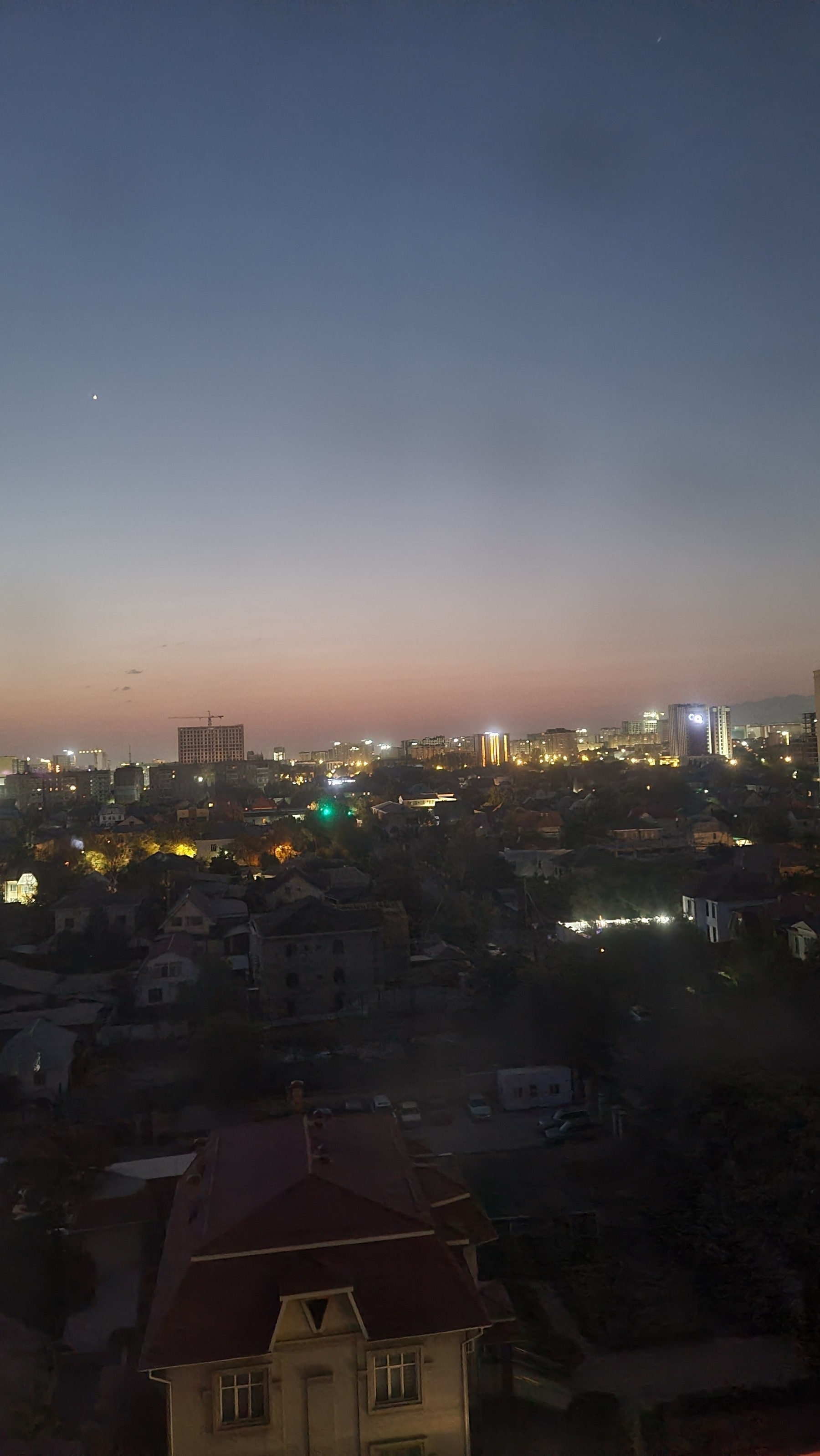 view of out a window at dawn. Buildings in the foreground are dark, but buildings in the background have lights