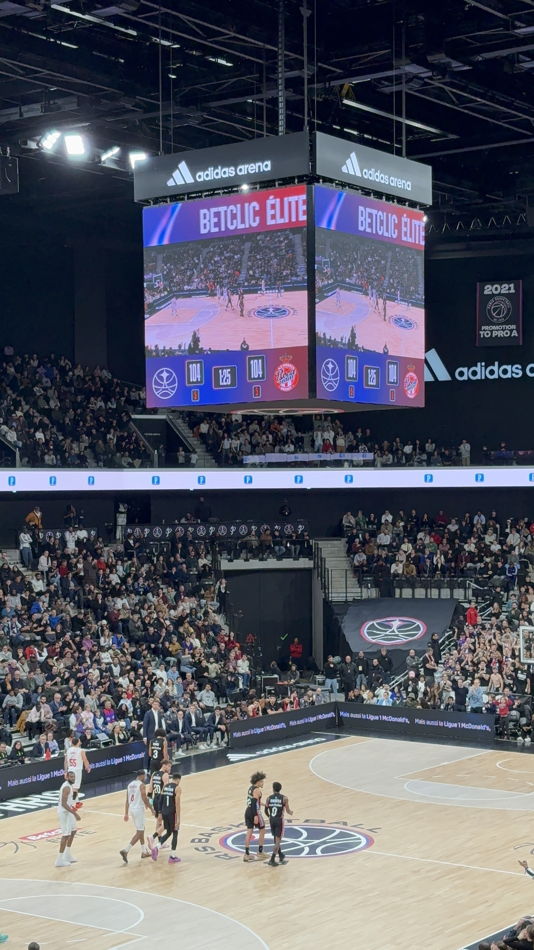 A basketball game is taking place at the adidas arena with a large, detailed scoreboard displaying the score and live action, surrounded by a crowd of spectators.