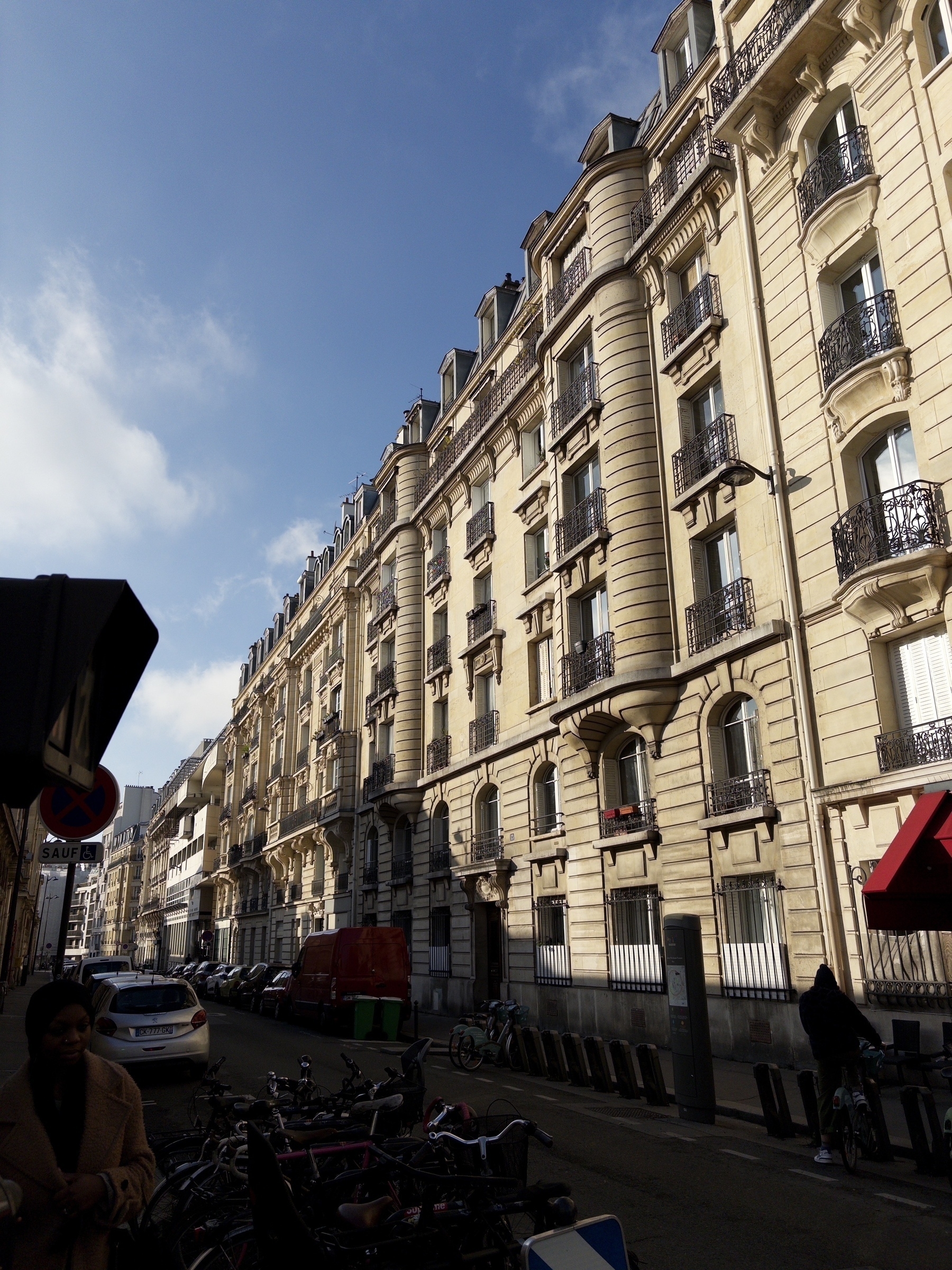 A street scene featuring classic European architecture with a row of ornate buildings, parked cars, bicycles, and pedestrians under a clear blue sky.