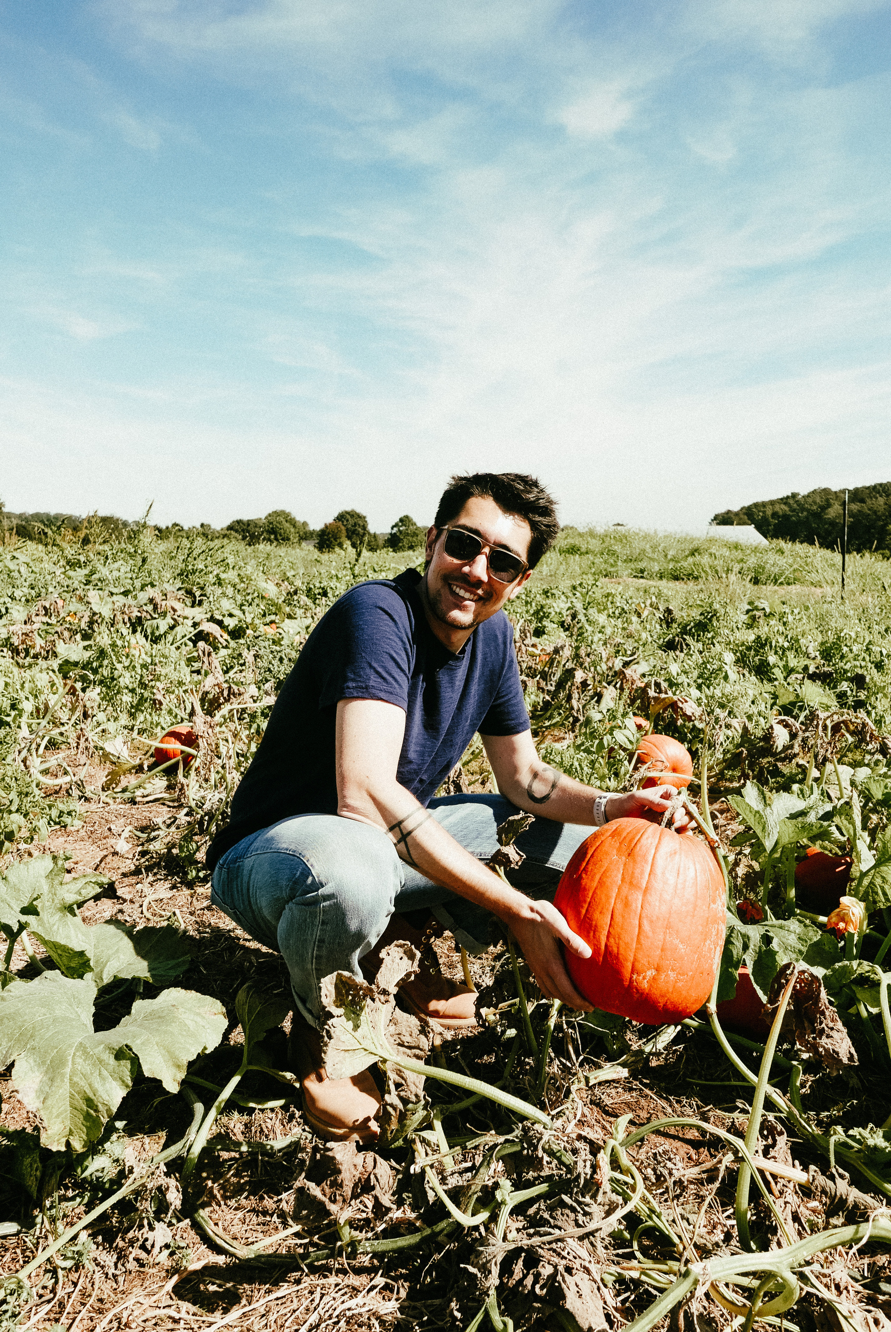 Auto-generated description: A person crouches in a pumpkin patch, holding a large orange pumpkin.