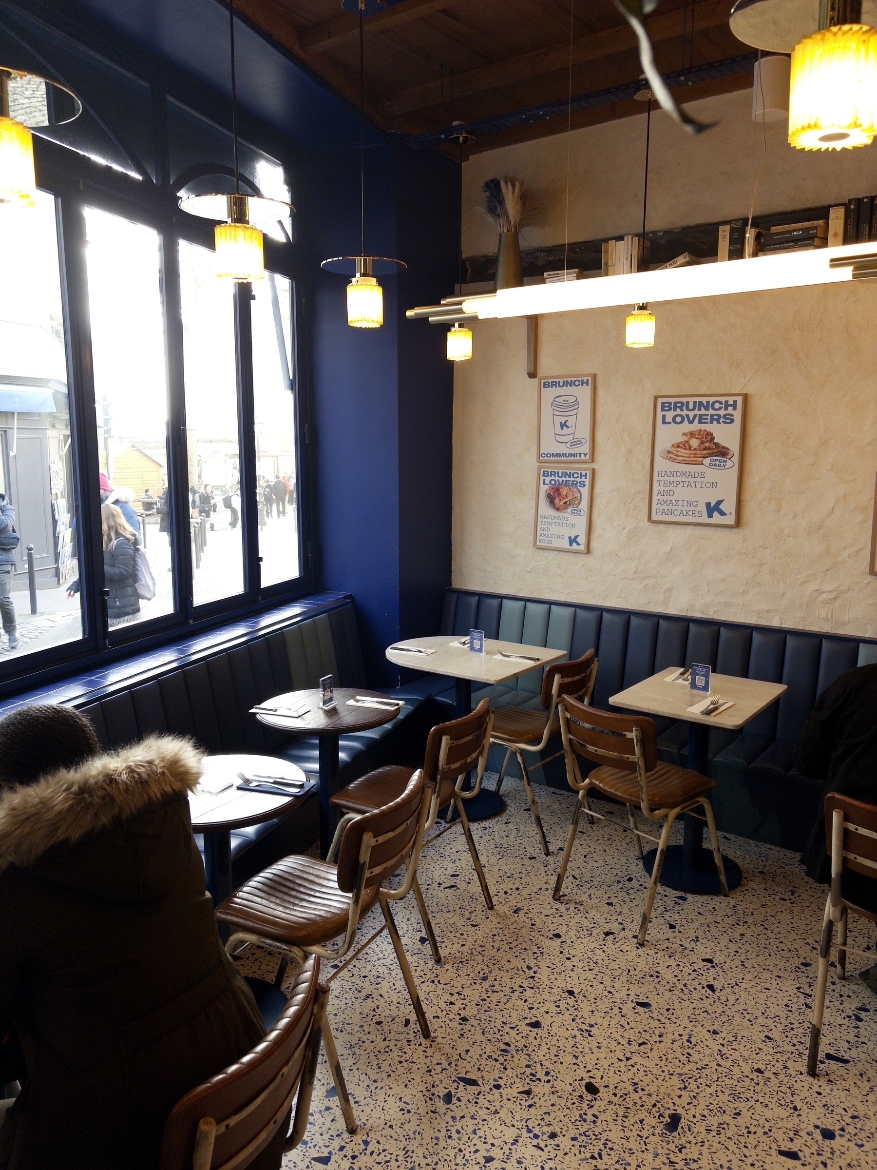 A cozy café interior features wooden chairs, tables, a cushioned bench lining the wall, and posters advertising brunch specials.