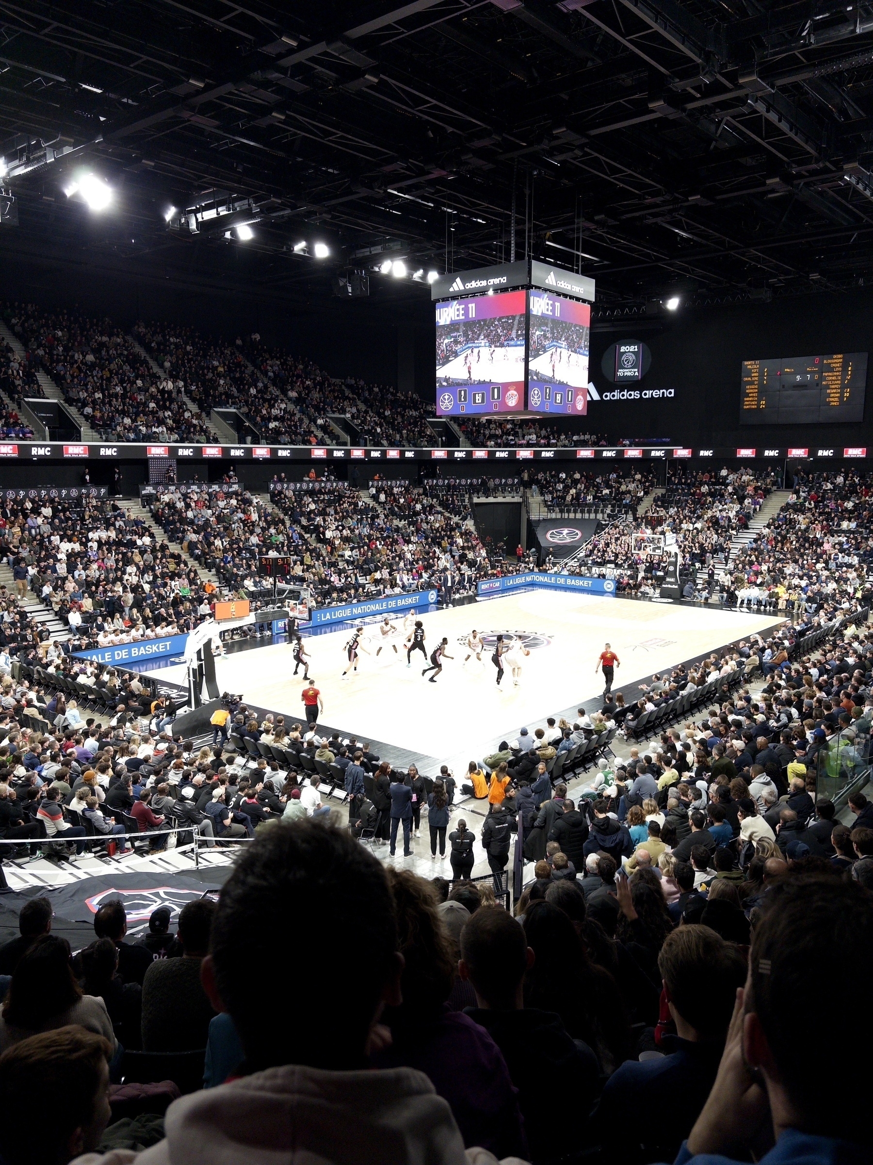 A packed indoor basketball arena features a game in progress, with a large audience watching from the stands.